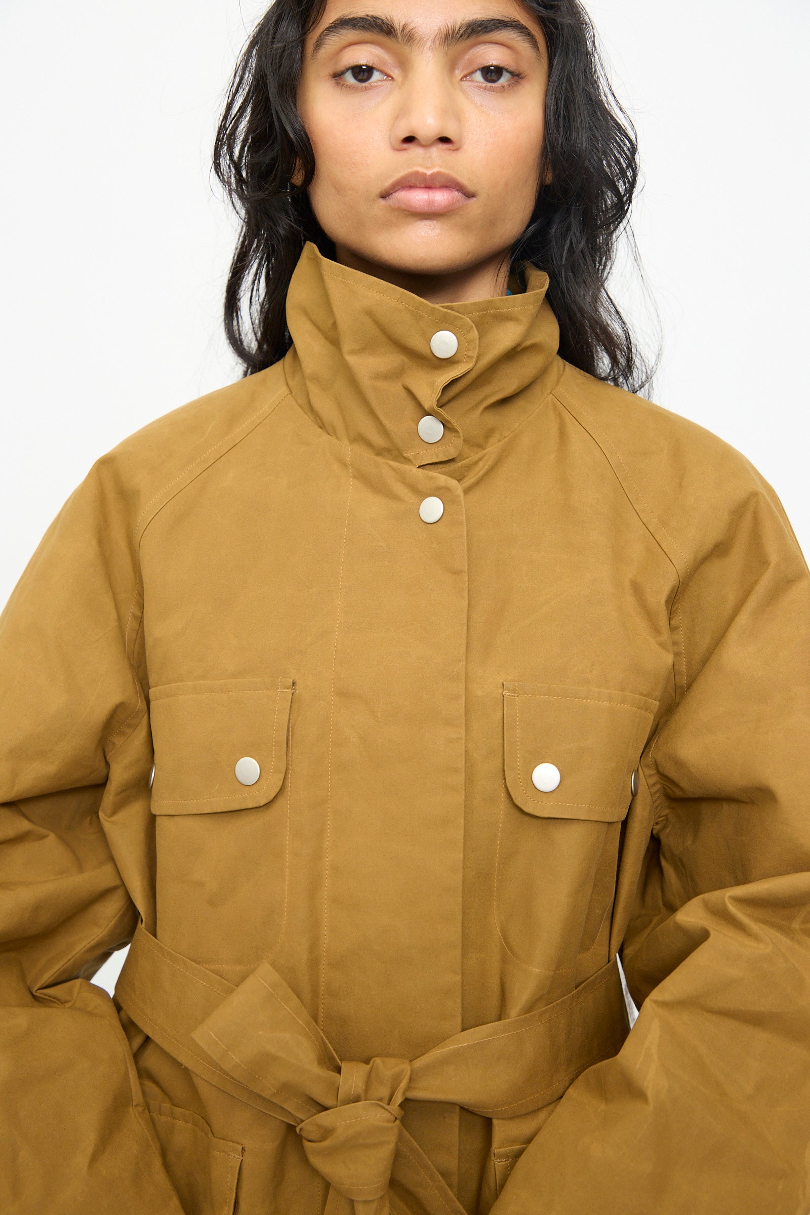 A person wearing the Olivia Cashmore Waxed Cotton Morison Parka in Ochre—featuring front pockets, a high collar, button closures, and a tied belt—stands against a plain light background.