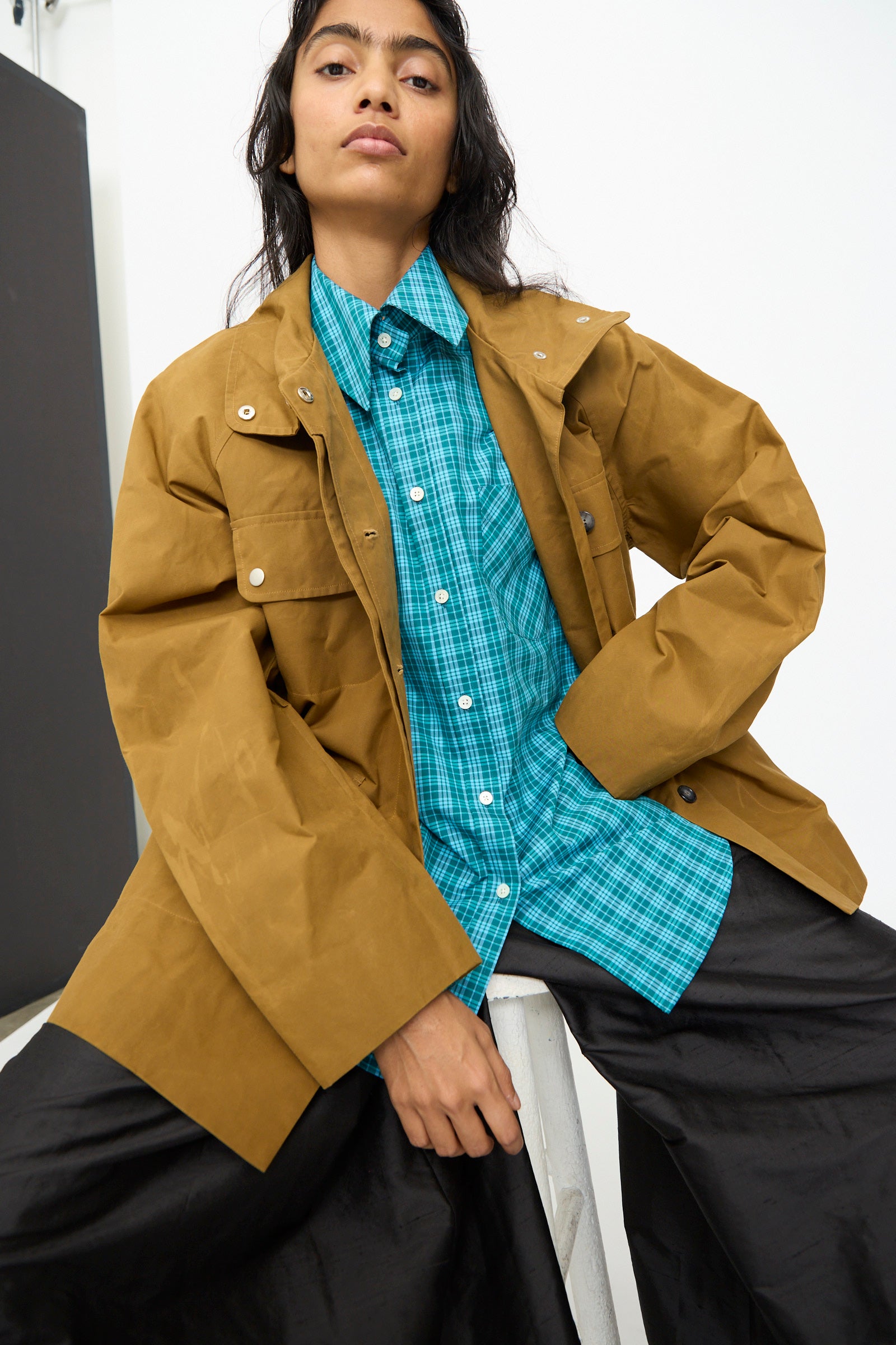 A person wearing the Waxed Cotton Morison Parka in Ochre by Olivia Cashmore, made in New Zealand, layered over a blue plaid shirt and black pants, sits on a white stool against a plain background.