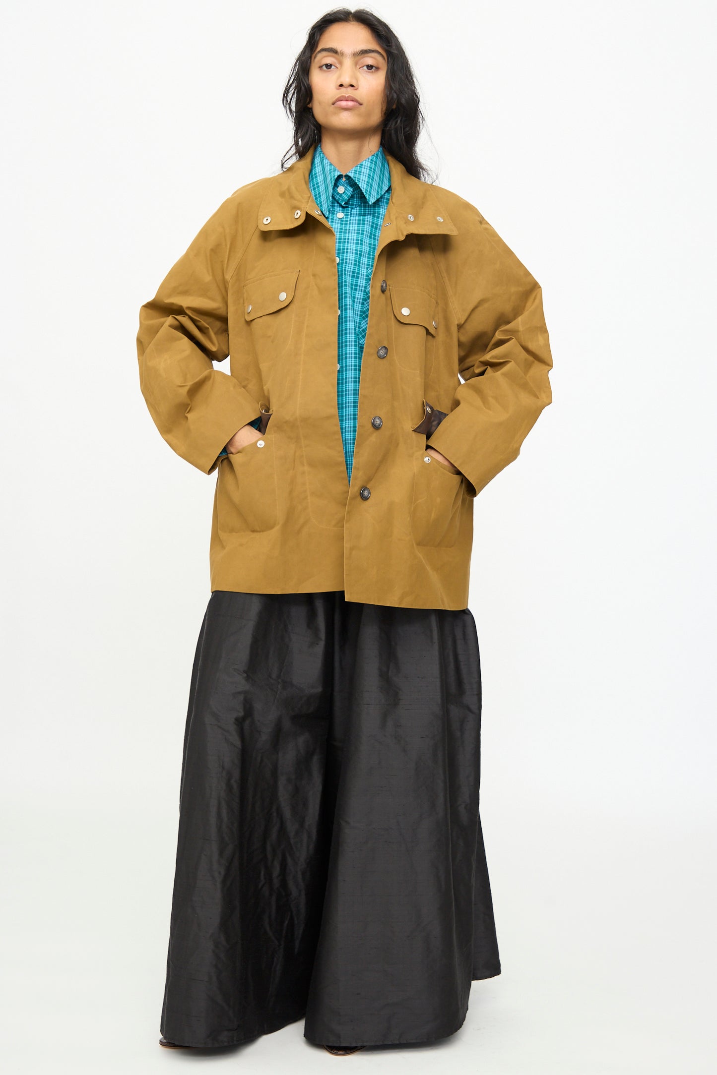 Wearing the Olivia Cashmore Waxed Cotton Morison Parka in Ochre over a blue plaid shirt and wide black pants, the person stands with hands in the jacket pockets against a plain white background.