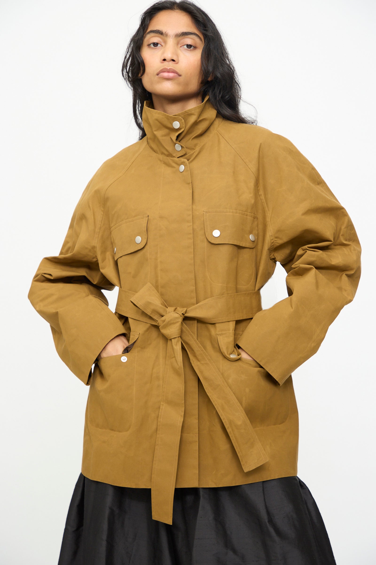 A person wearing the Olivia Cashmore Waxed Cotton Morison Parka in Ochre—a brown parka jacket with a belted waist, large front pockets, and silver buttons, crafted from durable waxed cotton—stands against a plain white background.