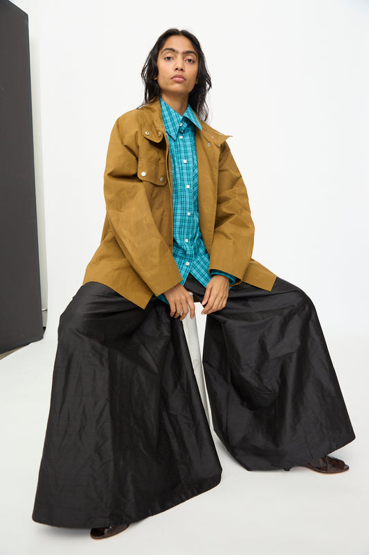 A person sits on a stool against a plain white background wearing the Olivia Cashmore Waxed Cotton Morison Parka in Ochre, belted at the waist and layered over a teal plaid shirt with wide black pants.