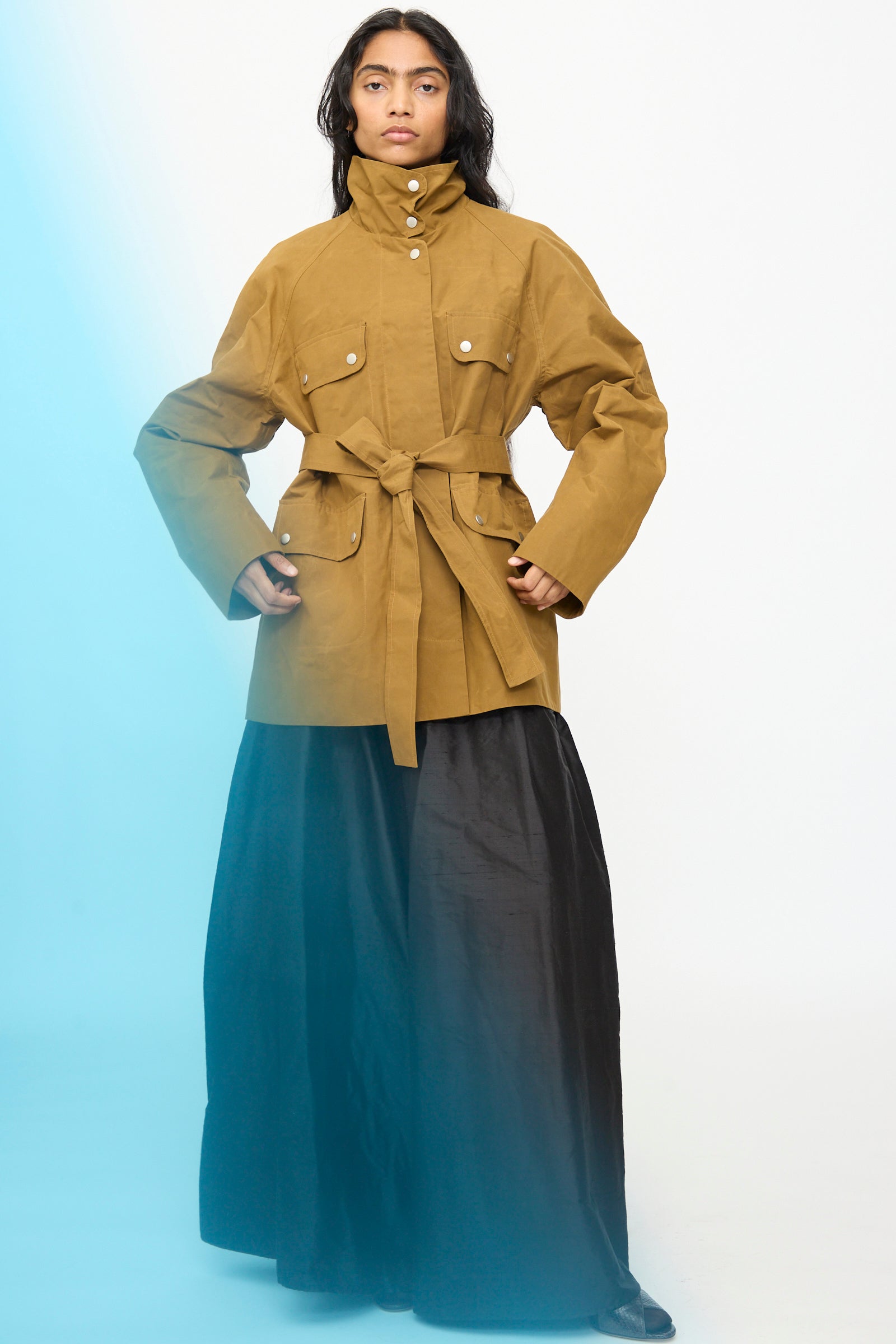 A person wearing the Olivia Cashmore Waxed Cotton Morison Parka in Ochre, featuring large pockets and a belted waist, paired with a long black skirt, stands against a plain light background with a subtle blue gradient on the left side.