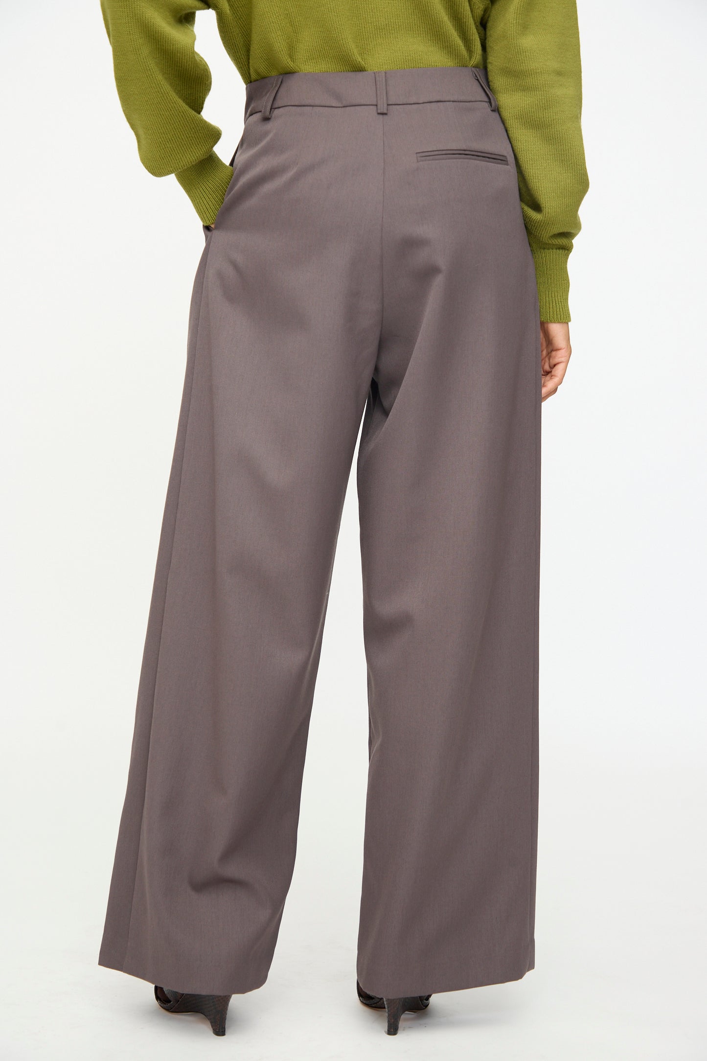 A person wearing the Olivia Cashmore Wool Marco Pant in Mud—a high-waisted, wide-leg tailored style—paired with a green long-sleeve top and heeled shoes, photographed from behind against a plain white background.