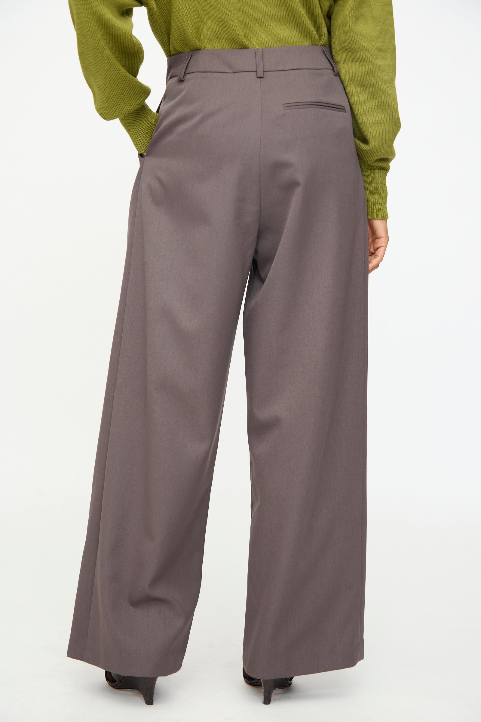 A person wearing the Olivia Cashmore Wool Marco Pant in Mud—a high-waisted, wide-leg tailored style—paired with a green long-sleeve top and heeled shoes, photographed from behind against a plain white background.