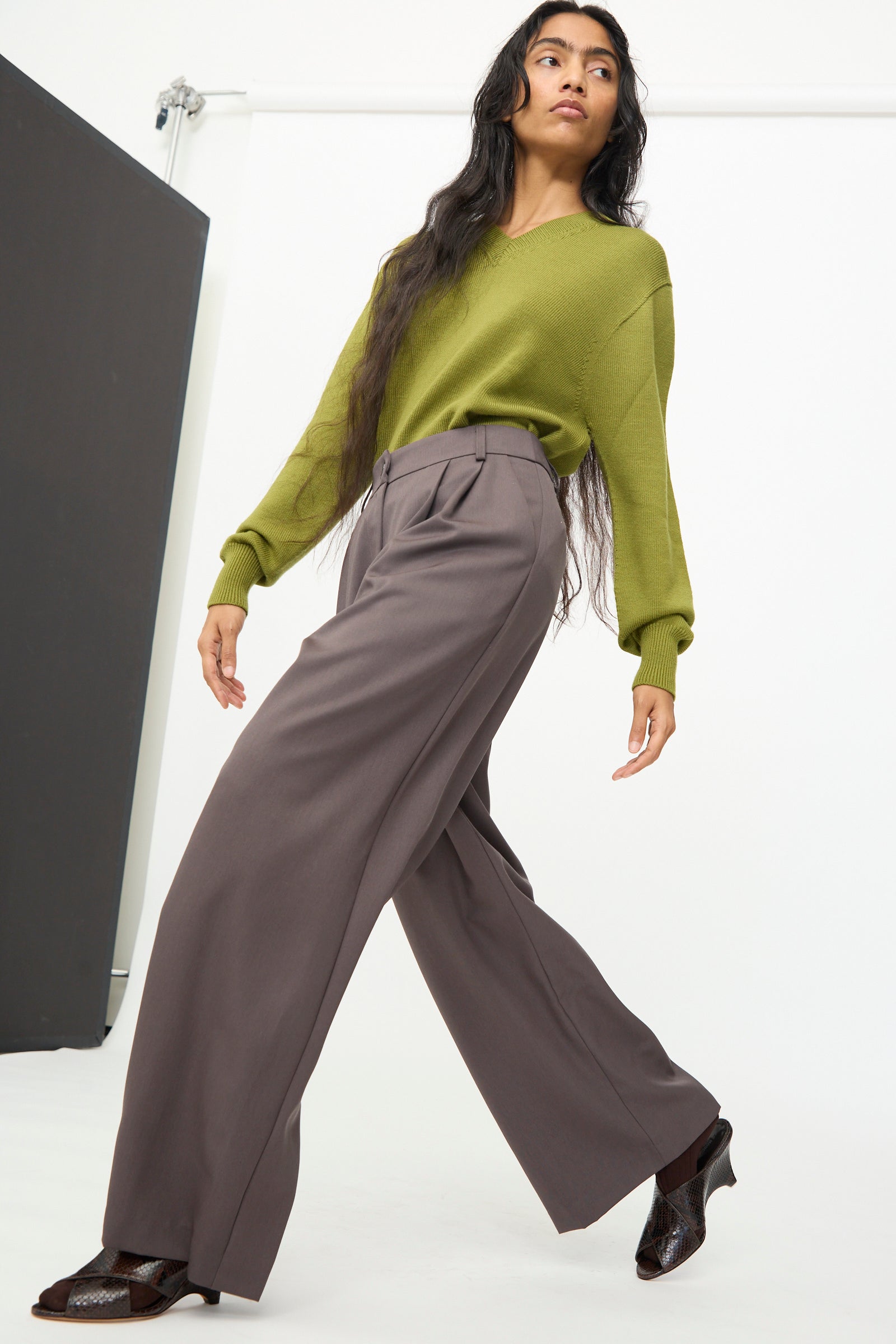 A person wearing a green sweater, Olivia Cashmore’s Wool Marco Pant in Mud, and brown shoes poses against a plain white background in a studio setting.