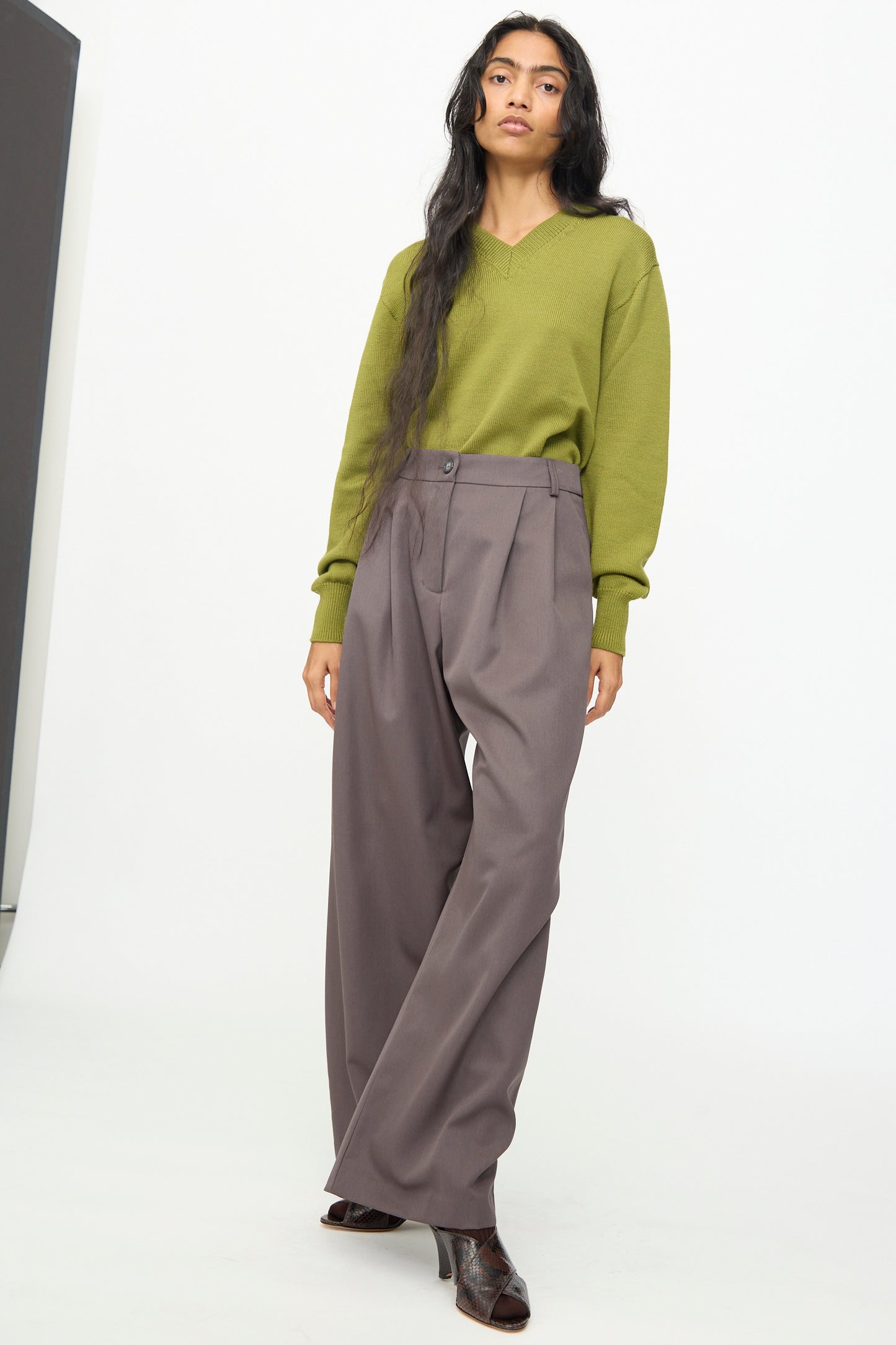 A woman stands against a plain background wearing a light green sweater, dark ankle boots, and Olivia Cashmore’s Wool Marco Pant in Mud—a pair of relaxed, tailored high-waist gray wool blend wide-leg pants.