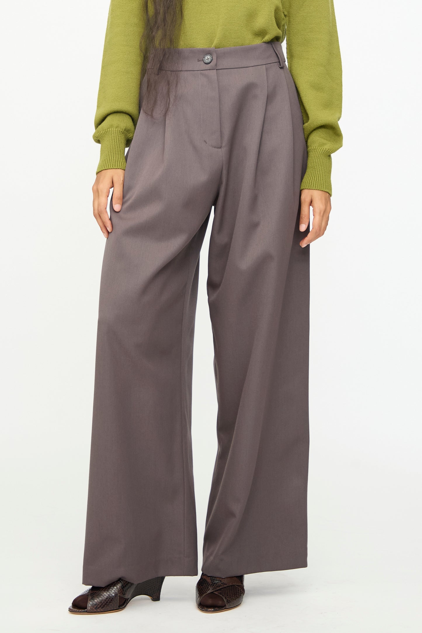 A person wearing the Olivia Cashmore Wool Marco Pant in Mud—a high-waist, wide-leg gray wool-blend trouser—paired with a green long-sleeve top and dark shoes, stands against a plain white background.