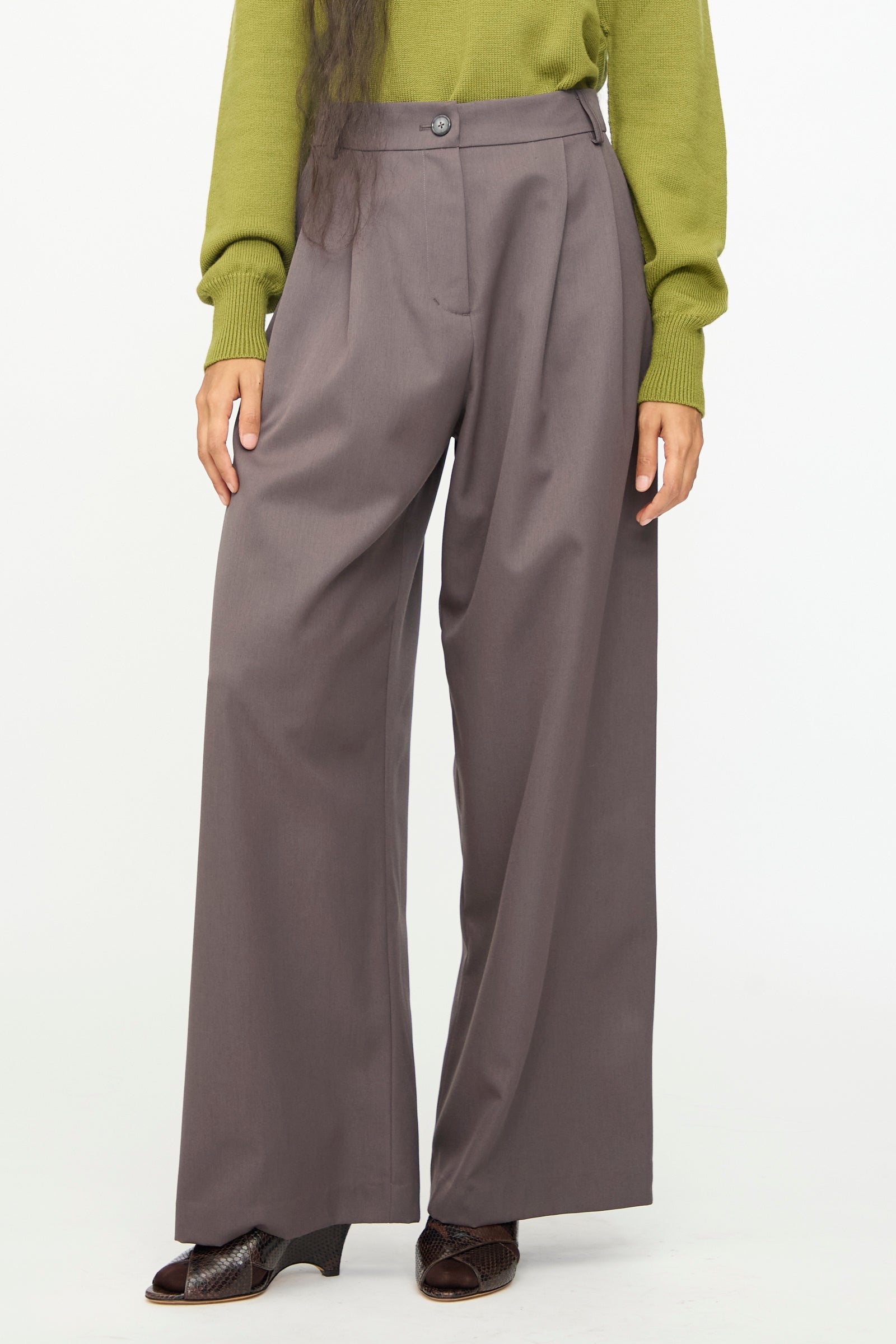 A person wearing the Olivia Cashmore Wool Marco Pant in Mud—a high-waist, wide-leg gray wool-blend trouser—paired with a green long-sleeve top and dark shoes, stands against a plain white background.
