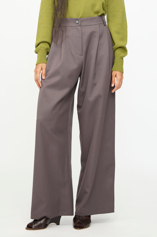 A person wearing the Olivia Cashmore Wool Marco Pant in Mud—a high-waist, wide-leg gray wool-blend trouser—paired with a green long-sleeve top and dark shoes, stands against a plain white background.