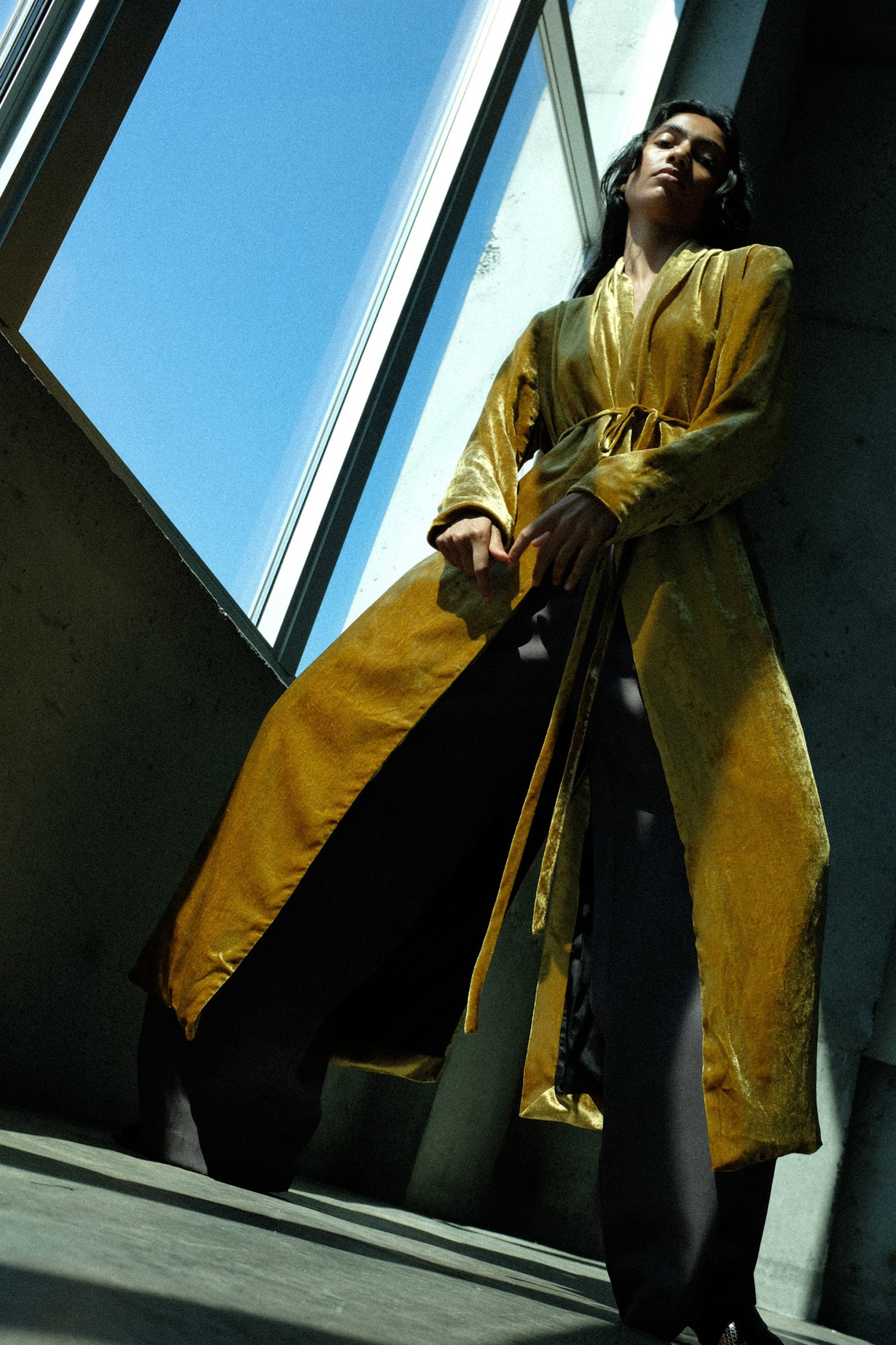 A model wearing the Olivia Cashmore Theresa Coat in Amber, standing in a modern concrete structure with large windows showing a blue sky.