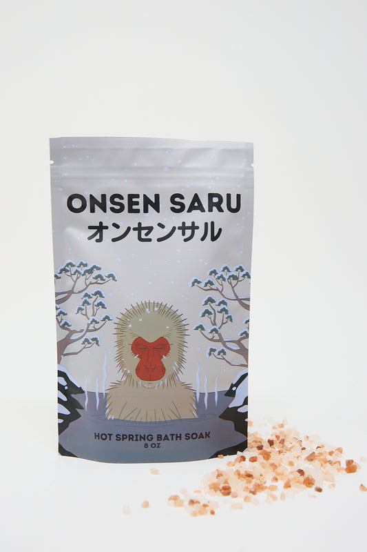 A bag of Onsen Saru hot spring bath soak with Japanese lettering and an illustration of a red-faced monkey in a snowy scene, accompanied by scattered Himalayan pink salt.