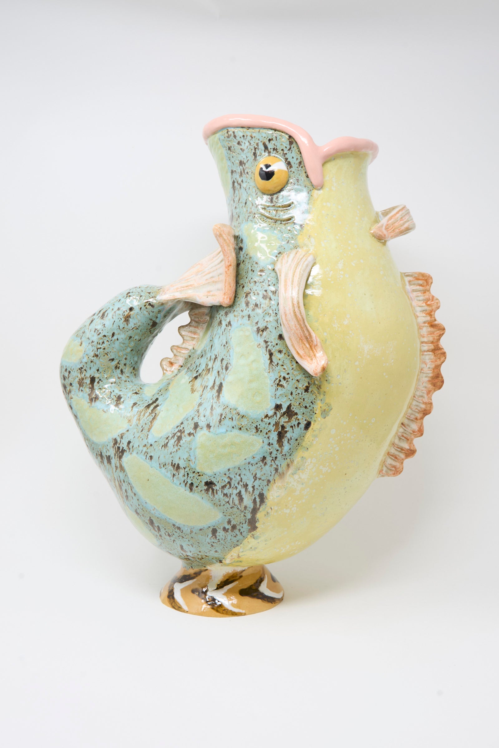 The Pearce Williams Fish Pitcher I is a ceramic, fish-shaped pitcher with a green and yellow glaze, textured fins, a fin-style handle, and a wide open mouth spout.