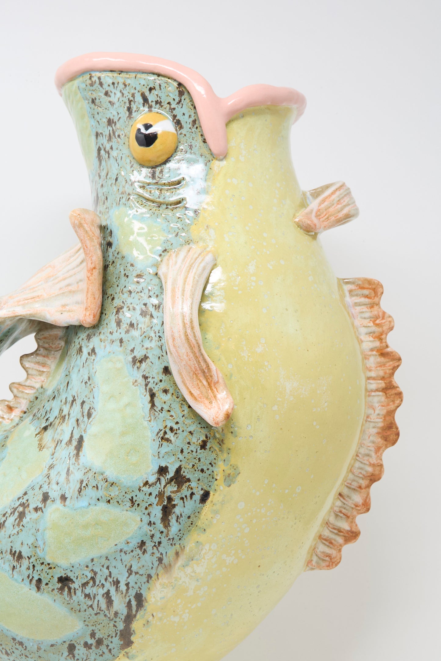 Close-up of the Fish Pitcher I by Pearce Williams, a handmade stoneware vase shaped like a fish with glossy yellow, green, and blue hues, textured fins, and a pink-edged mouth set against a plain white background.