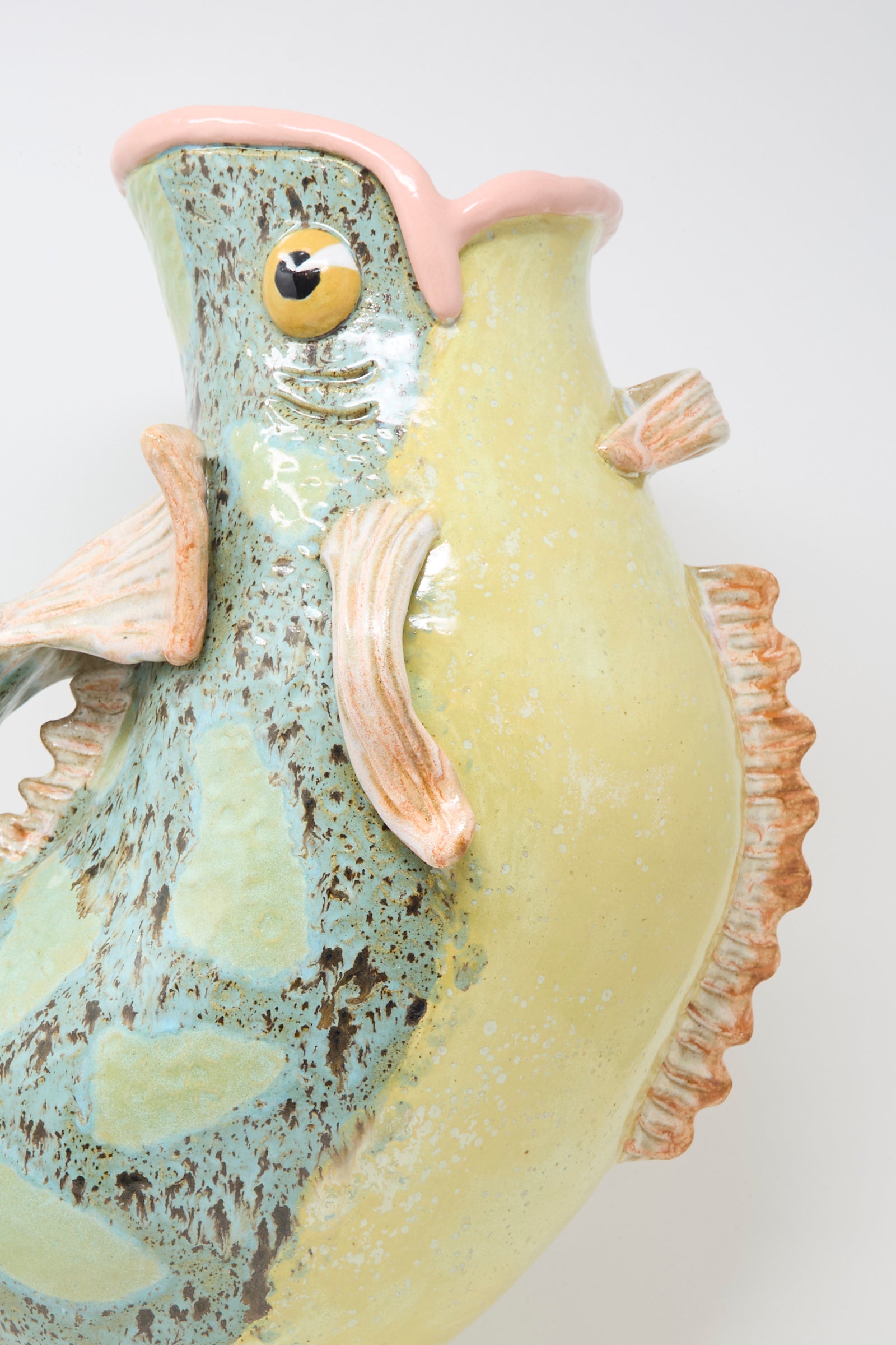 Close-up of the Fish Pitcher I by Pearce Williams, a handmade stoneware vase shaped like a fish with glossy yellow, green, and blue hues, textured fins, and a pink-edged mouth set against a plain white background.