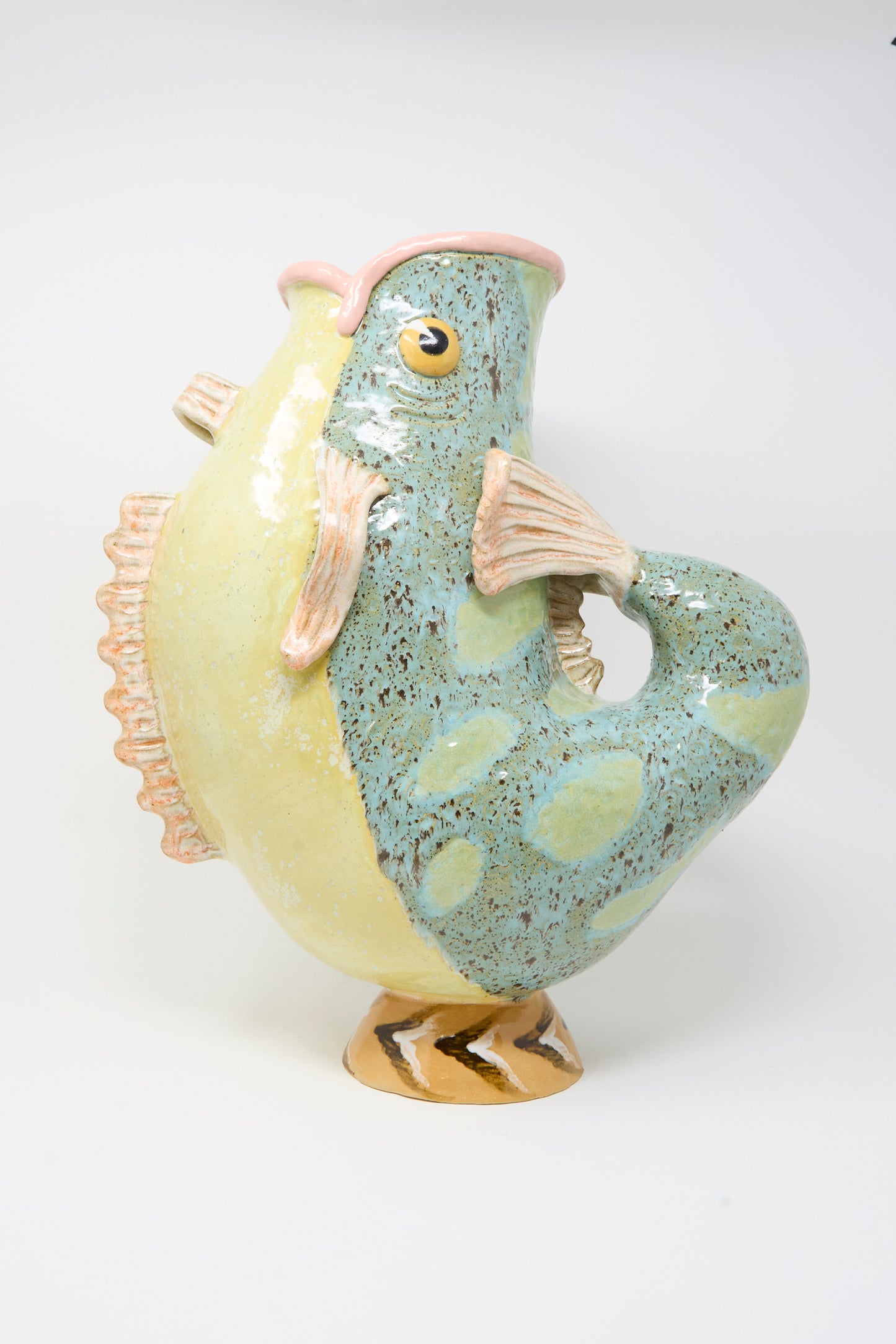 The Fish Pitcher II by Pearce Williams is a ceramic, fish-shaped vase featuring a curved tail handle and vibrant yellow, blue-green, and pink hues—a playful decorative piece to brighten any space.