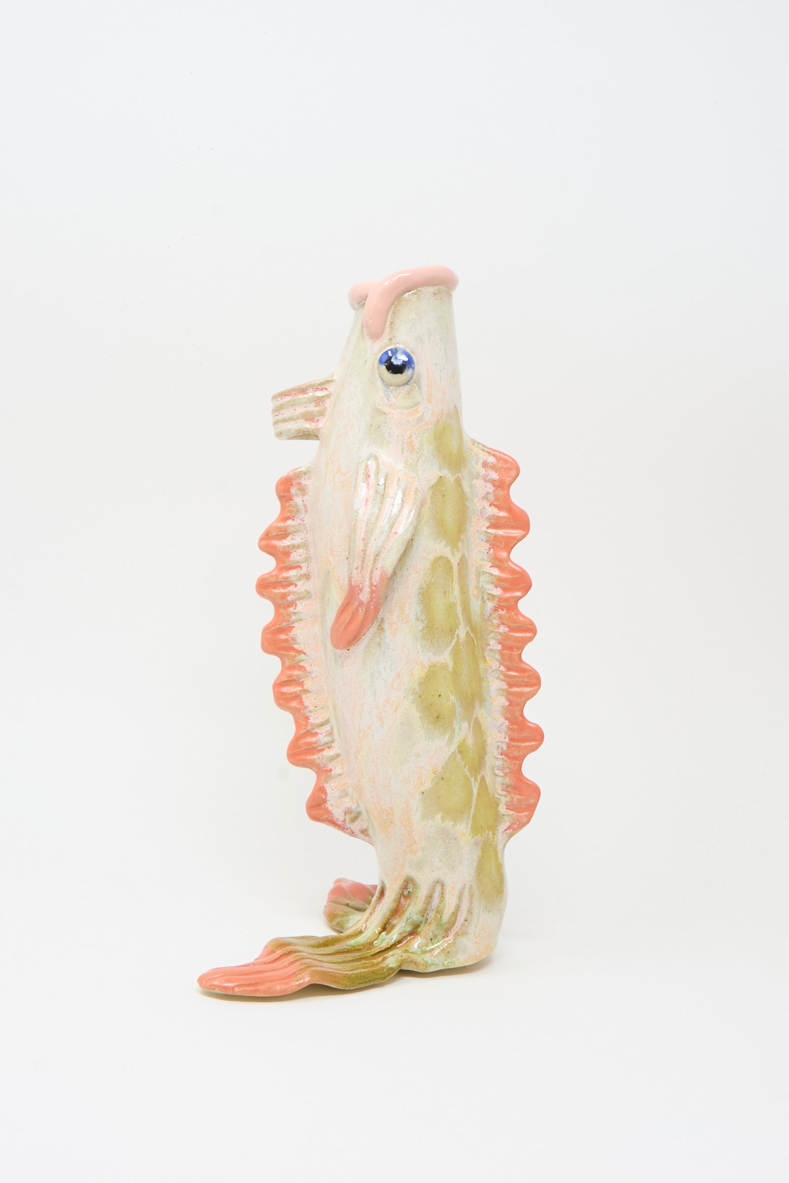 The Fish Vase in Coral II by Pearce Williams is a handmade ceramic sculpture from New Orleans, featuring a pale body with greenish spots, orange fins, and a glossy blue eye, standing upright against a plain white background.
