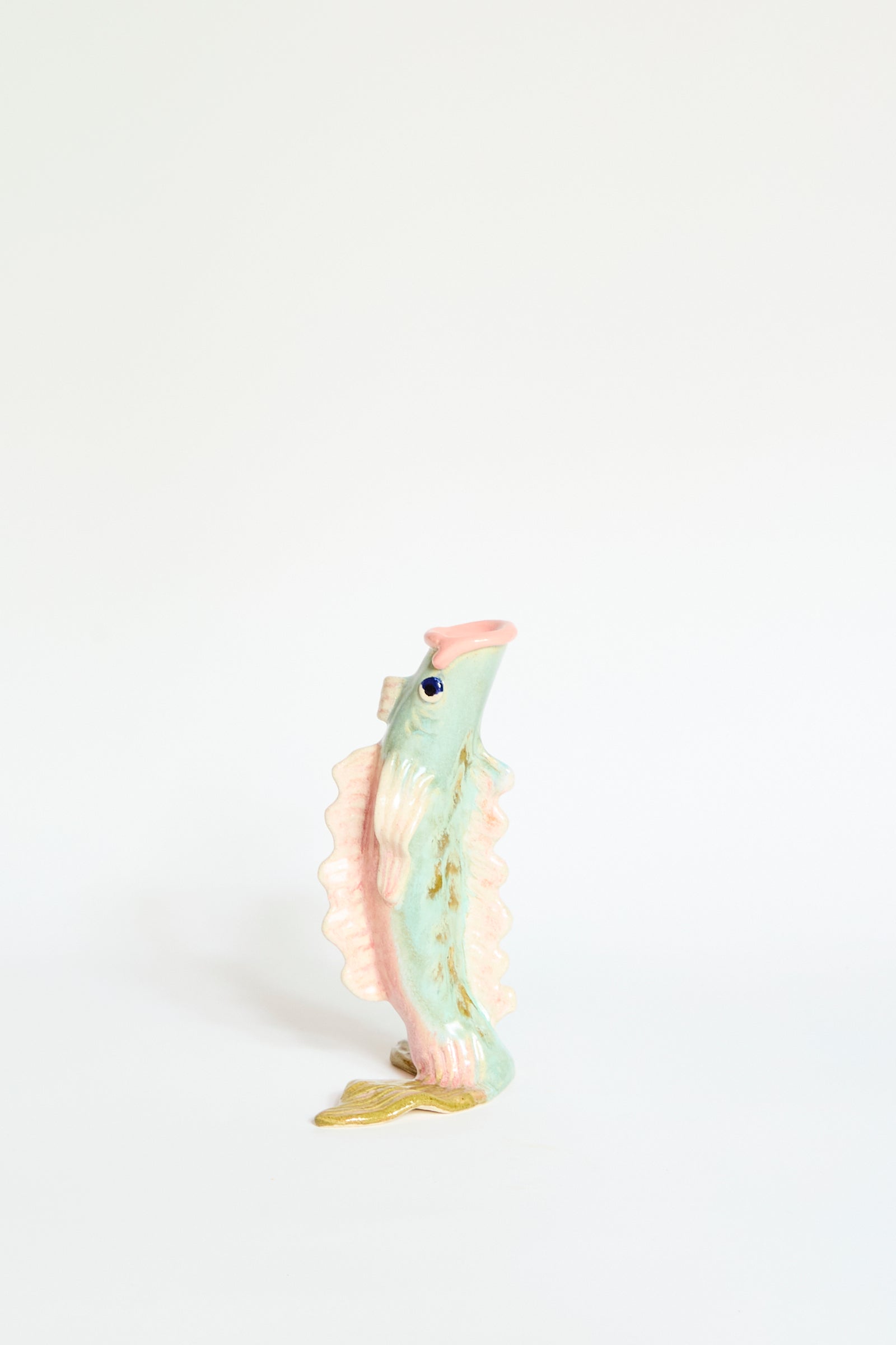 The Pearce Williams Fish Vase Small in Green is a handmade, pastel fish-shaped vase with blue eyes and a golden tail fin, inspired by New Orleans style, shown upright against a plain white background.