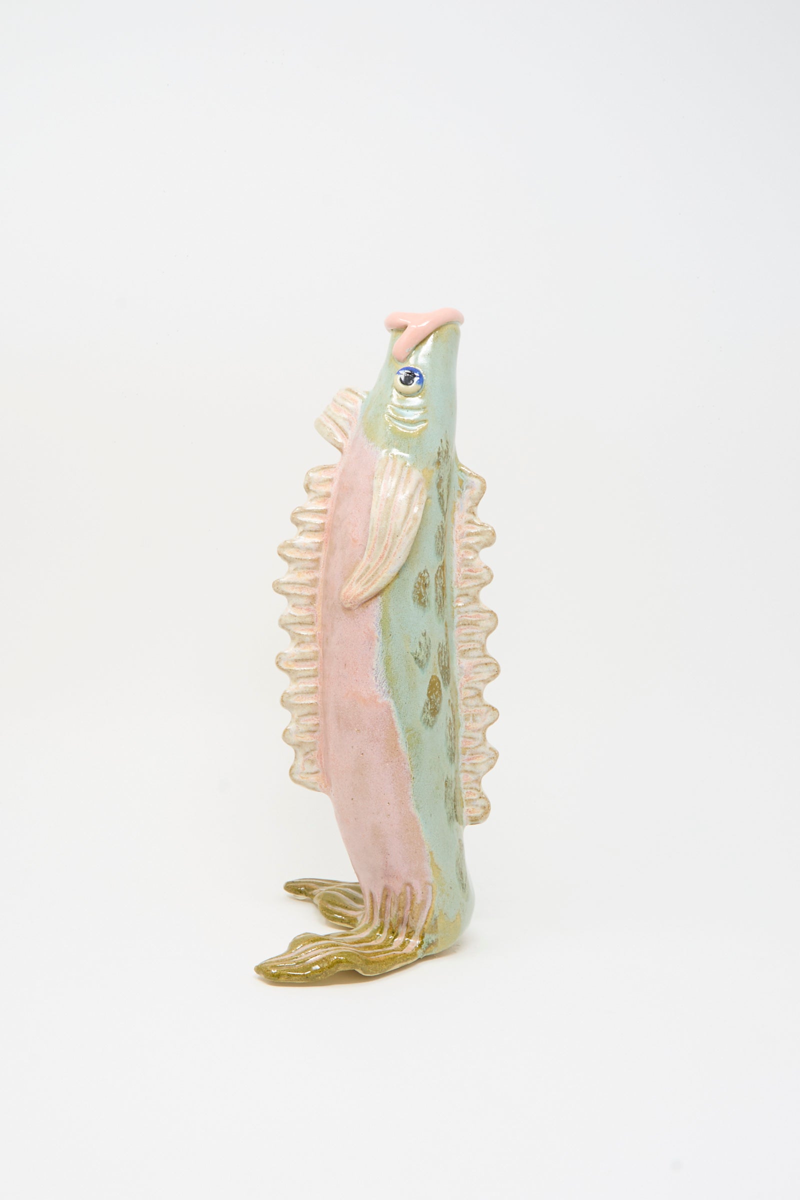The Pearce Williams Fish Vase in Green is a handmade stoneware upright vase shaped like a fish, with pastel green, pink, and gold hues and detailed fins and scales, shown on a plain white background.