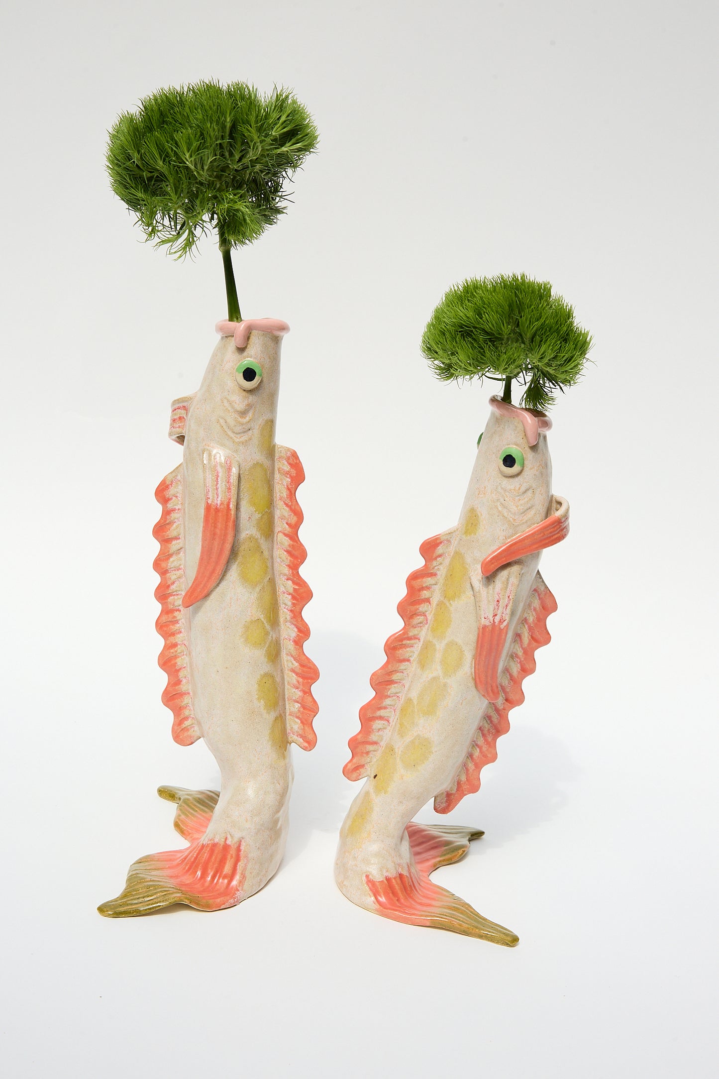 Two whimsical glazed stoneware koi fish sculptures, each adorned with a lush green plant on top, standing upright on their tail fins against a white background. The Pearce Williams Fish Vase in Pink.