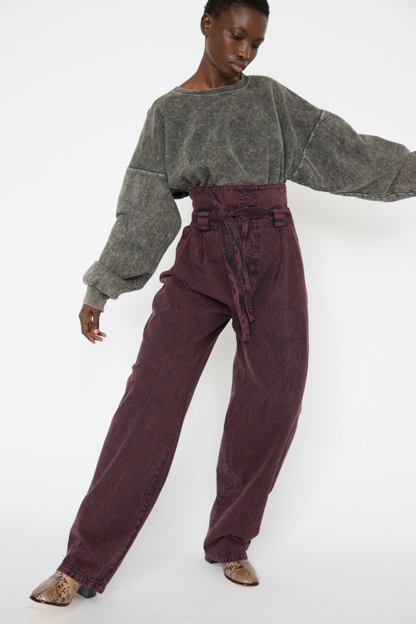 A woman in a grey oversized sweater and Rachel Comey's Acid Madrona Denim Bowman Pant in Boisonberry poses against a plain background and looks down.