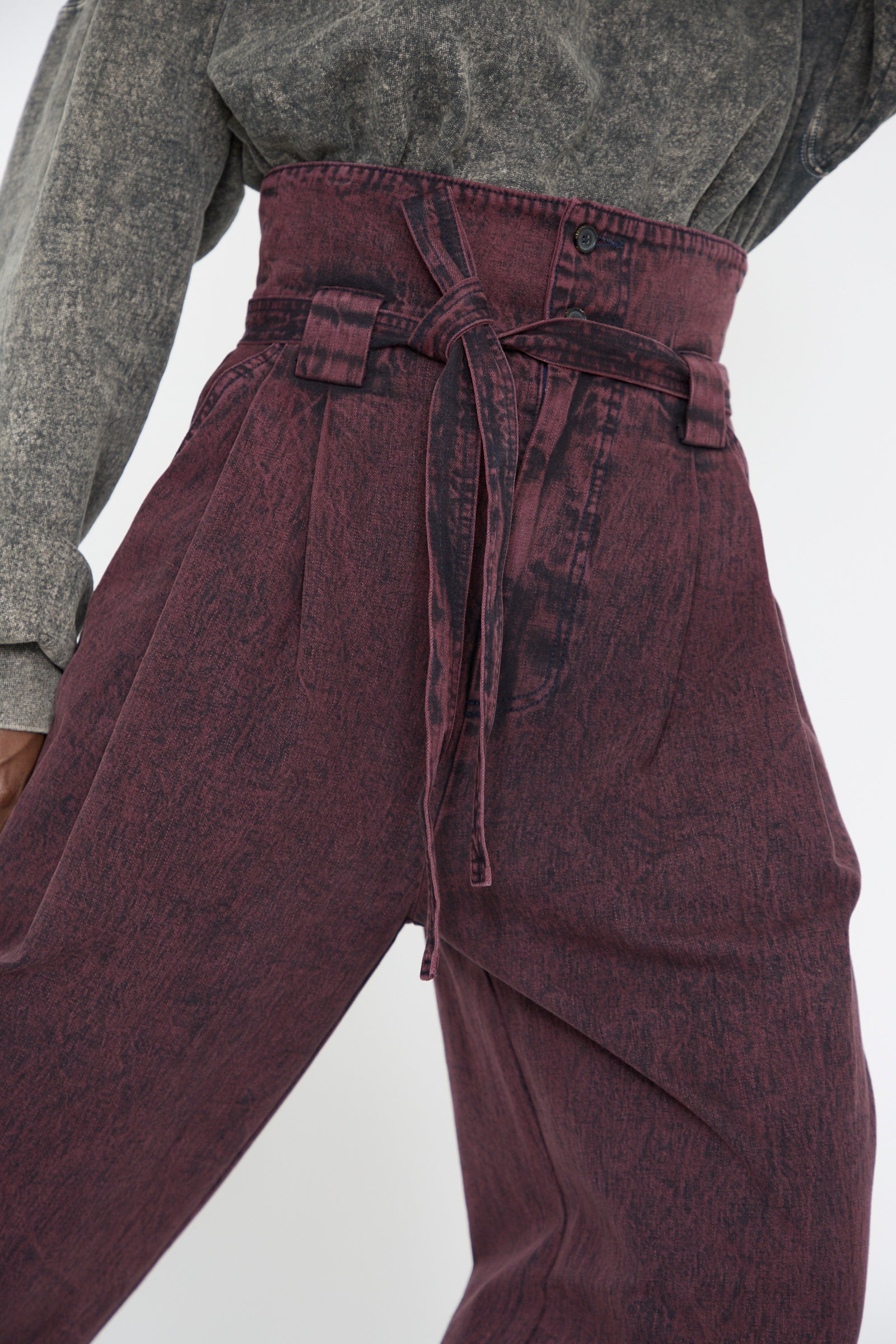 A close up of a woman wearing the Rachel Comey Acid Madrona Denim Bowman Pant in Boisonberry, featuring a high waist, wide legs, and a self-tie belt, paired with a gray shirt.