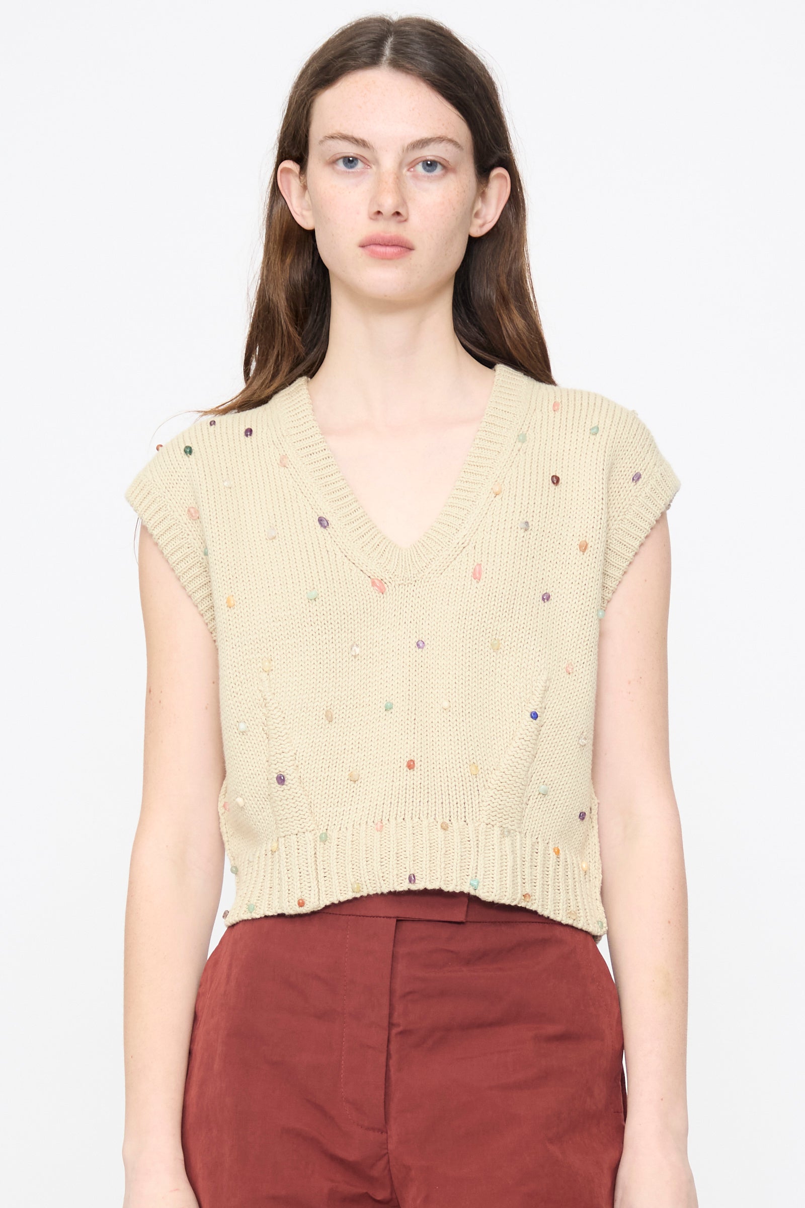 A woman wears the Acrea Top in Dove by Rachel Comey—a beige, chunky knit vest with colorful dots—paired with high-waisted maroon pants, standing against a plain white background.