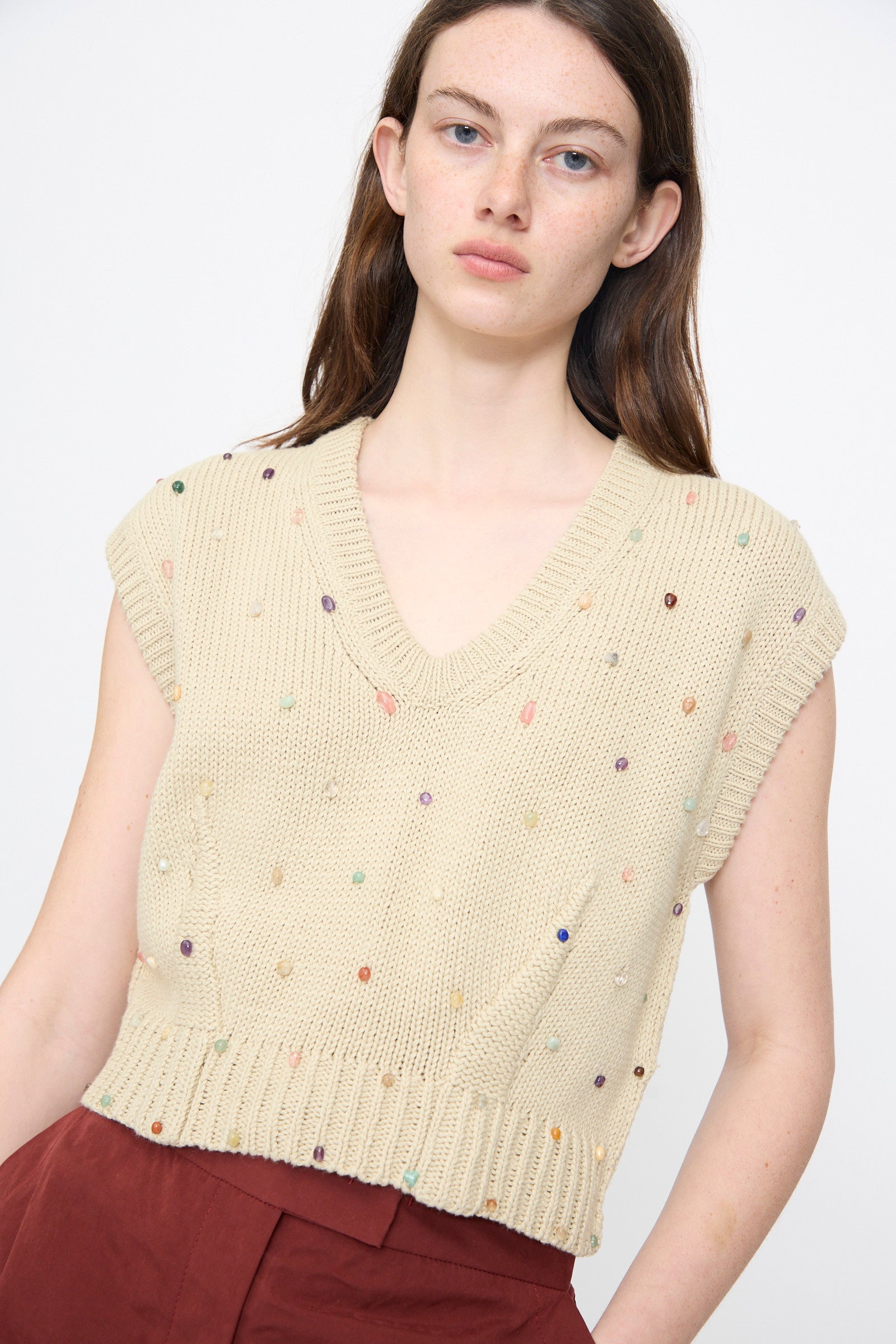 A woman wearing the Rachel Comey Acrea Top in Dove, a chunky knit beige sleeveless sweater with small multicolored dots, and high-waisted maroon pants stands against a plain light background.