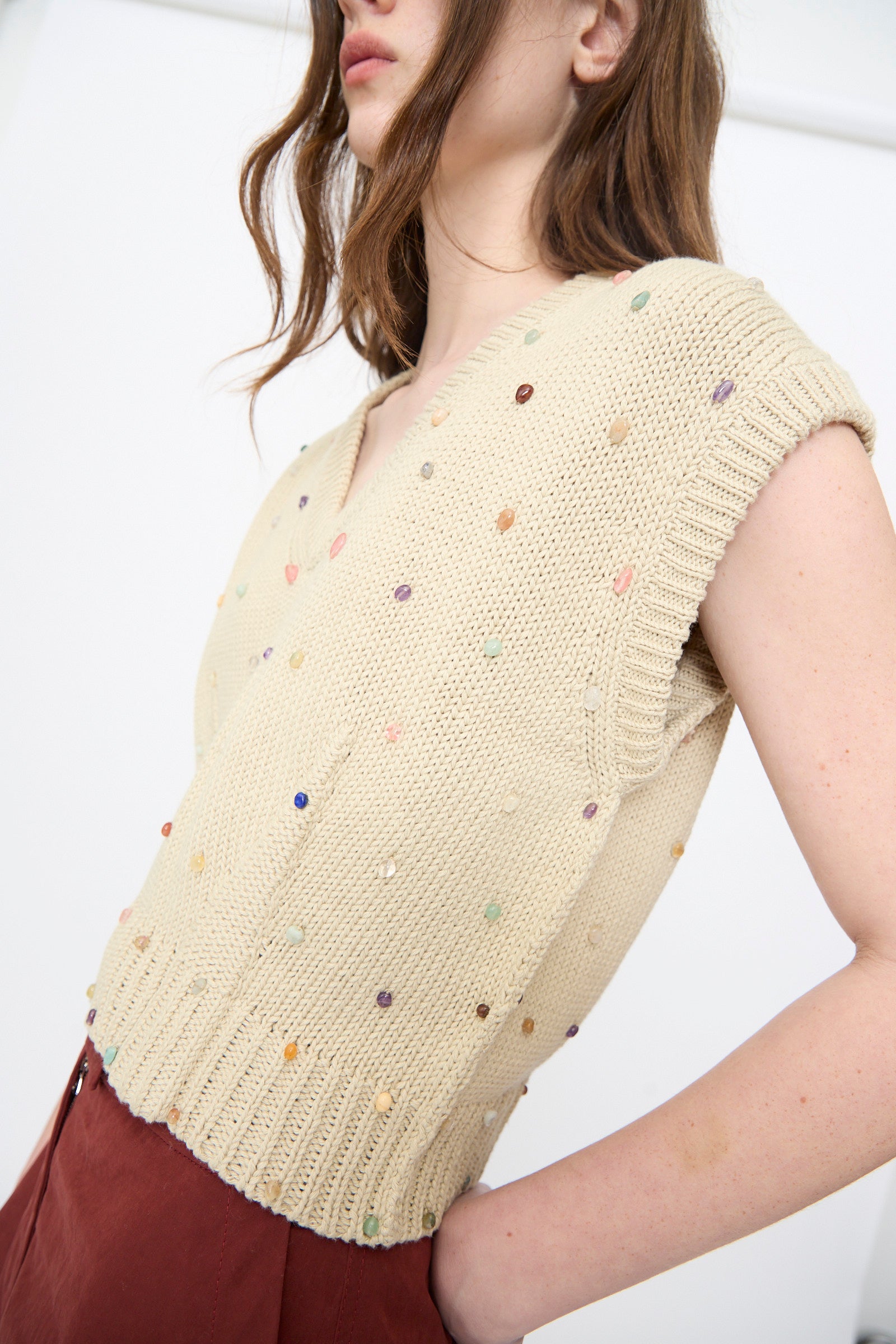 A person wears the Rachel Comey Acrea Top in Dove—a beige, short-sleeved chunky knit vest with colorful small beads—paired with high-waisted burgundy trousers, standing against a plain white background.