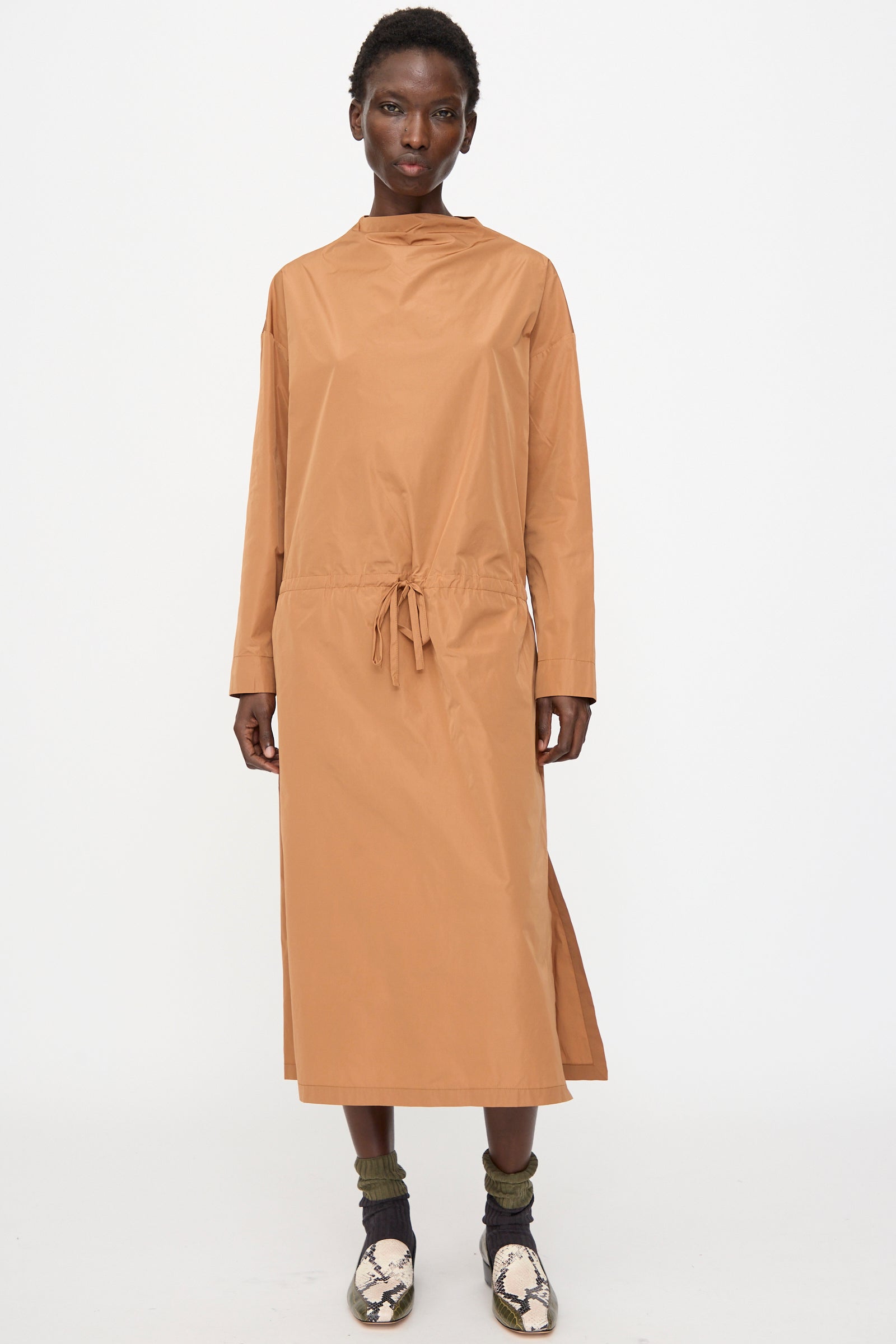 A person faces forward in the Rachel Comey Alberry Dress in Copper, a loose, long sleeve, water-resistant taffeta midi dress with a drawstring waist, styled with green socks and snakeskin shoes against a plain background.