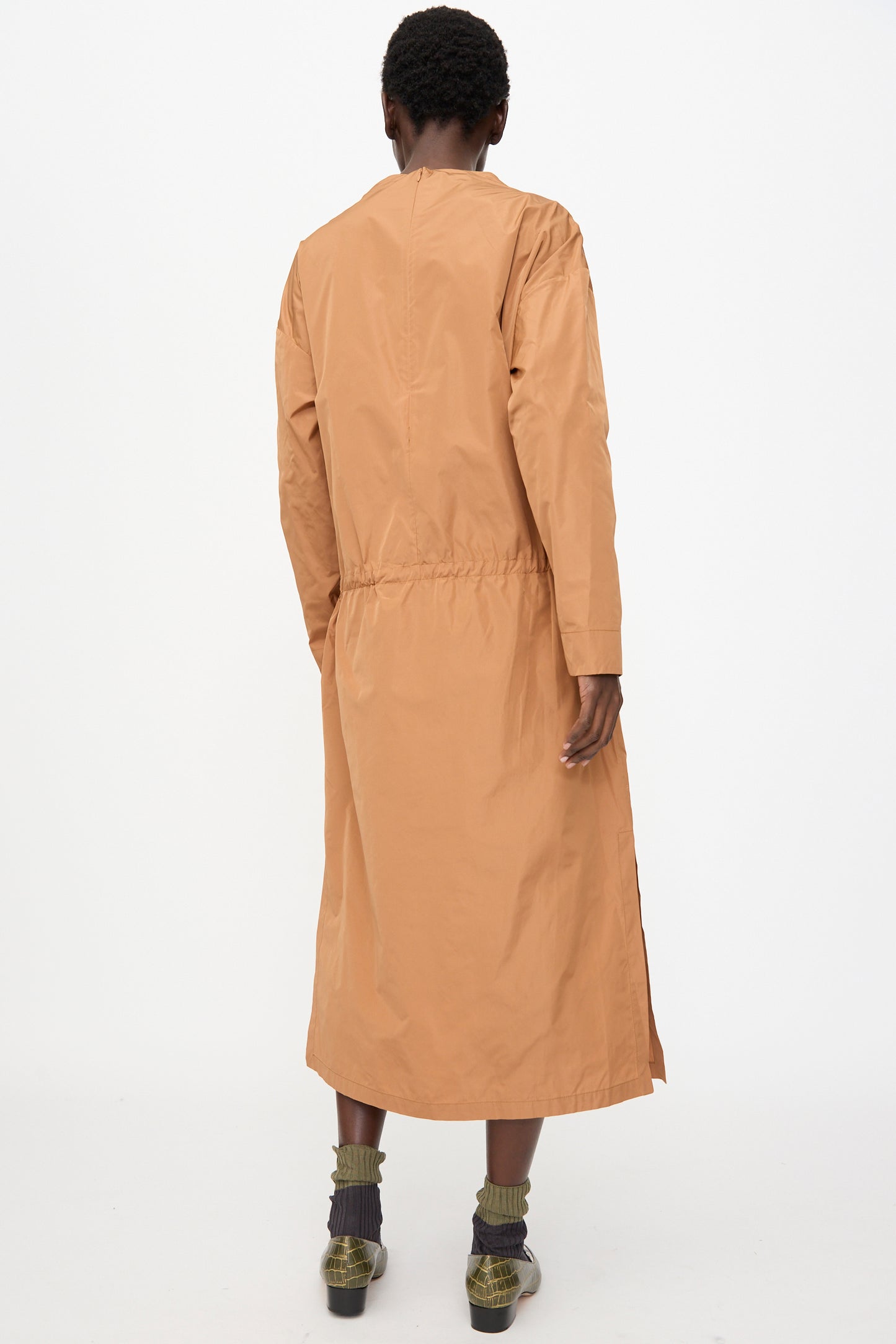 A person stands facing away, wearing the Rachel Comey Alberry Dress in Copper, paired with gray socks and green shoes, against a plain white background.