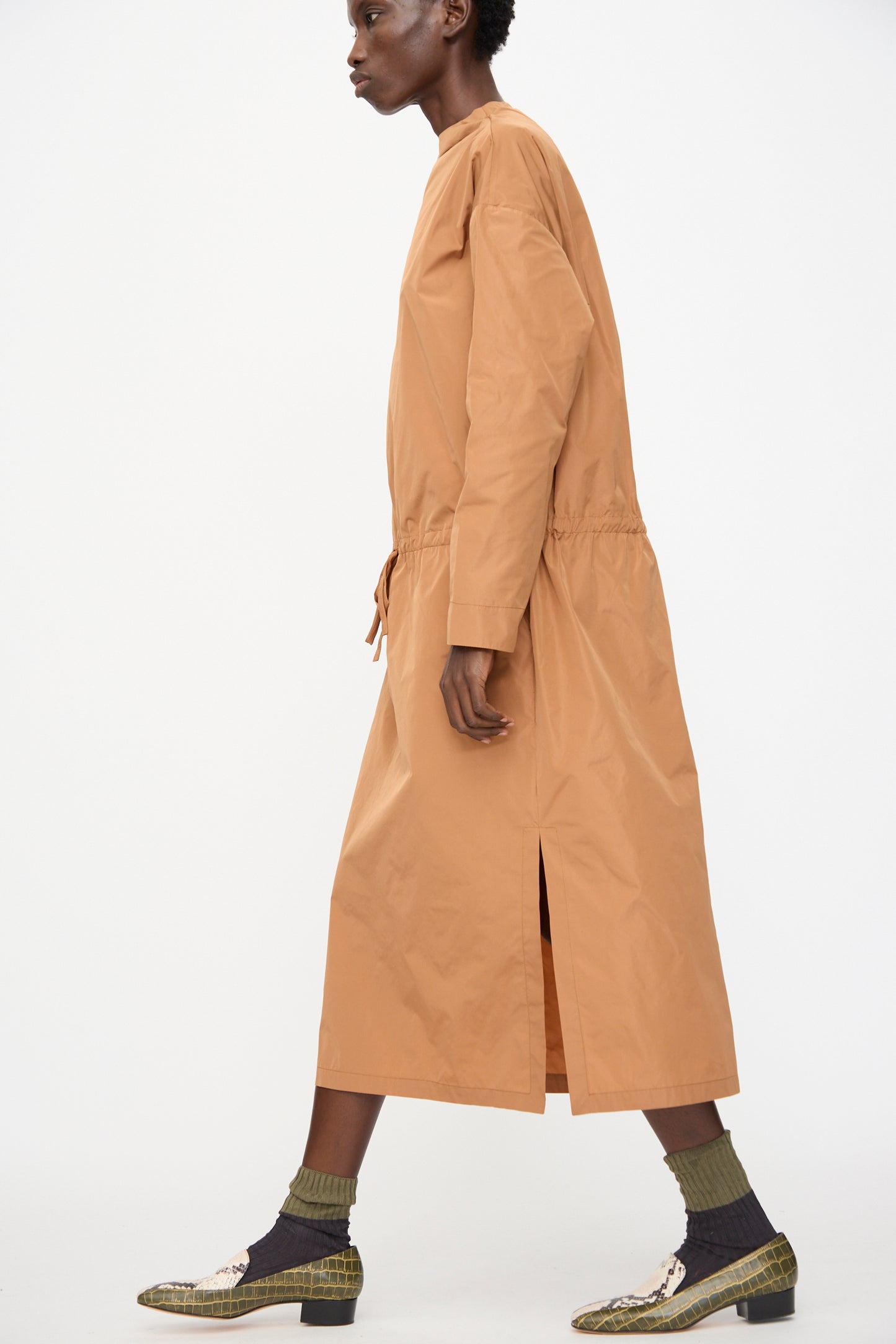 Wearing the Rachel Comey Alberry Dress in Copper—a tan recycled polyester, long sleeve midi dress with a drawstring waist—paired with patterned heels and green socks, a model walks against a plain white background.