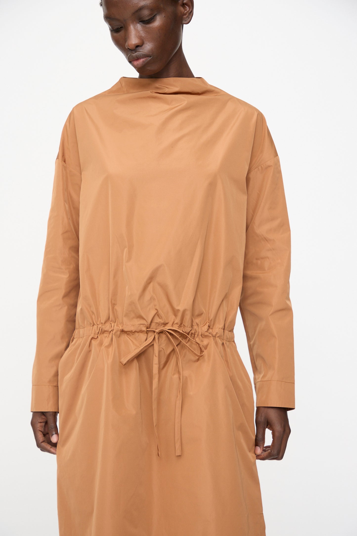 A person wears the Rachel Comey Alberry Dress in Copper, a long sleeve midi dress made from recycled polyester with a cinched waist, standing against a plain white background.