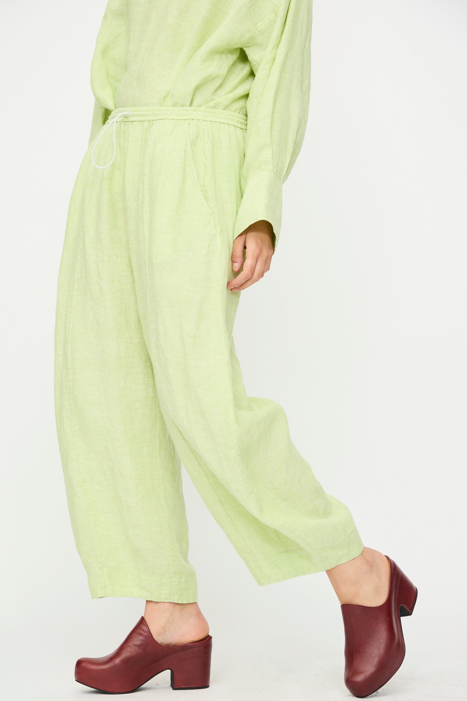 A person wears the Rachel Comey Ashbolt Pant in White Sage—light green, textured, wide-leg with an elastic waistband—paired with burgundy platform clogs, standing against a white backdrop.