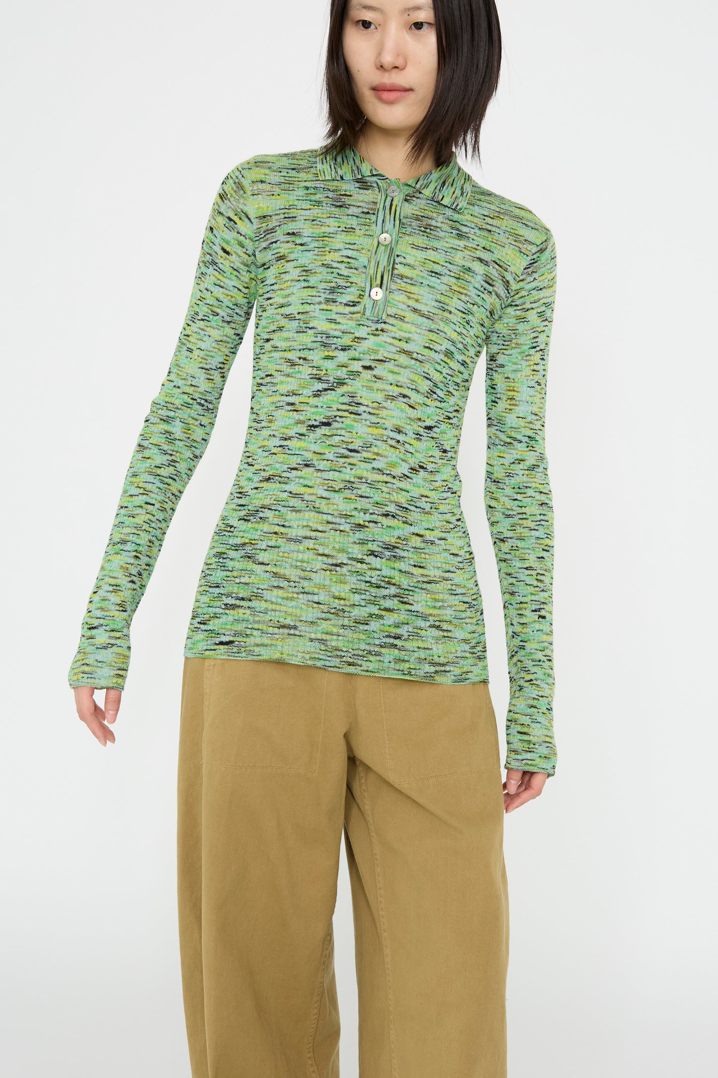 A person dressed in the Rachel Comey Avril Polo in Lime Multi Spacedye long-sleeve sweater and light brown pants stands against a crisp white background.