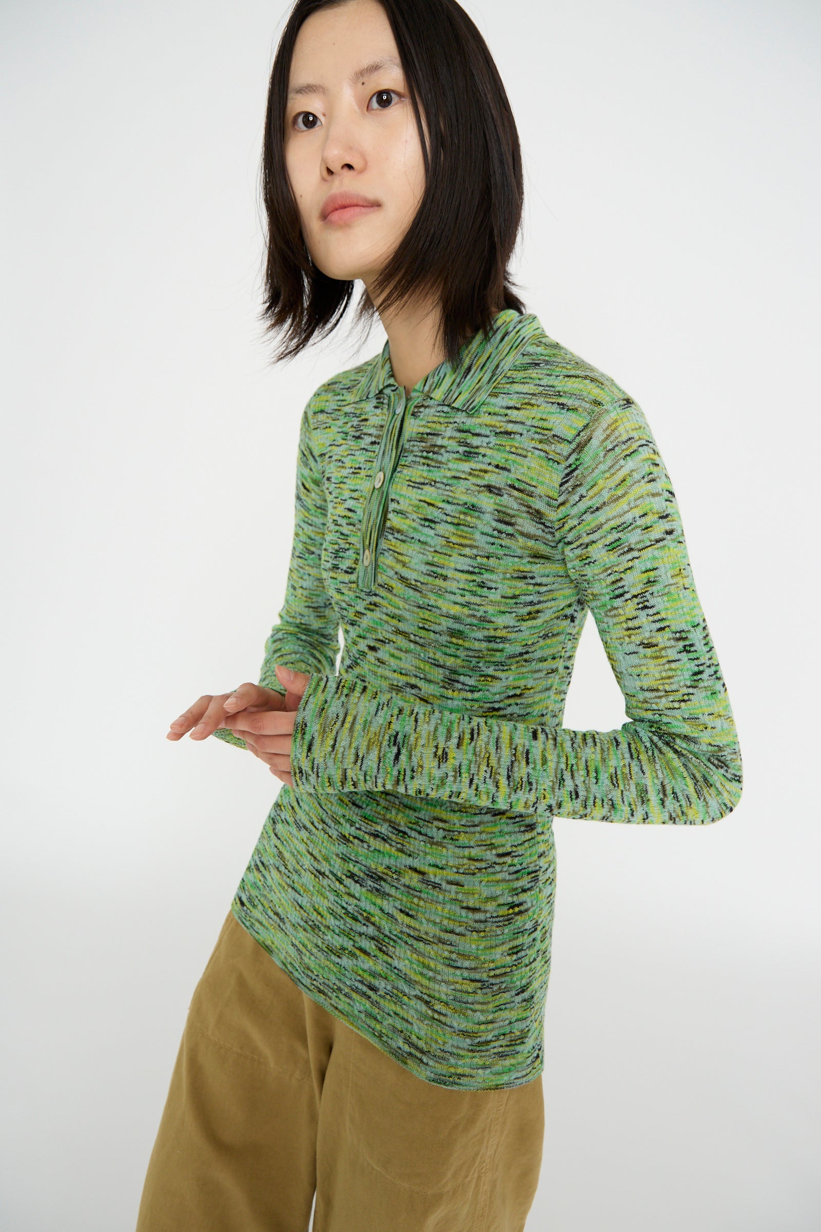 A person with long hair stands against a plain background, wearing the Avril Polo in Lime Multi Spacedye by Rachel Comey and beige pants.