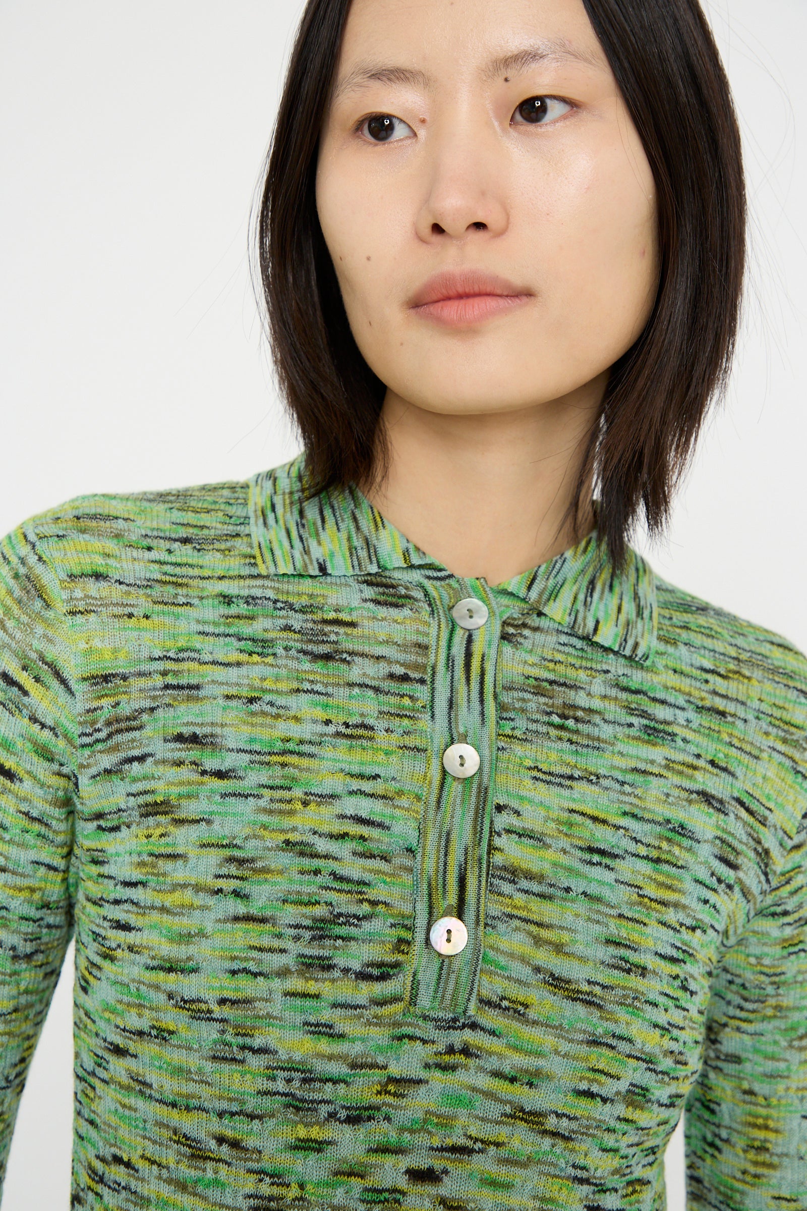 A model with straight, shoulder-length hair is wearing the Avril Polo in Lime Multi Spacedye by Rachel Comey, a green and black patterned collared shirt with buttons against a white backdrop. 