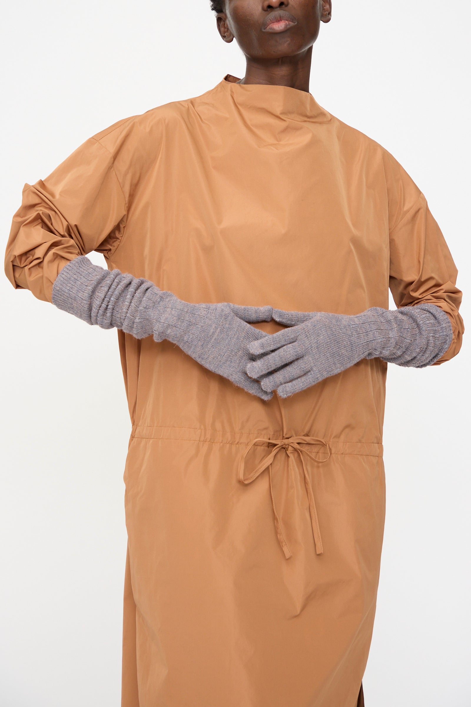 Person wearing a loose, tan long-sleeve dress with a tie at the waist and Rachel Comey Baalam Gloves in Dusty Mauve, made in Peru, standing against a plain white background.
