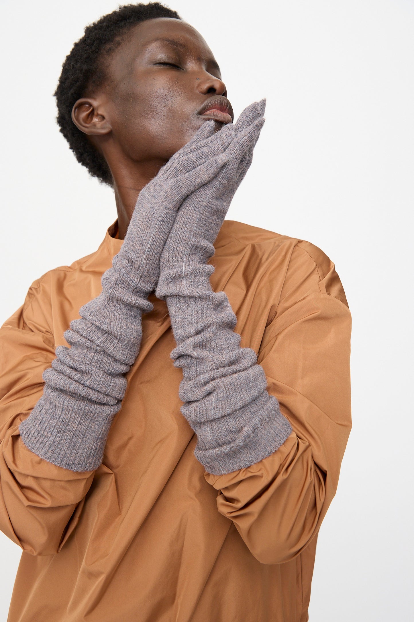 A person wearing a light brown top and Rachel Comey Baalam Gloves in Dusty Mauve touches their face with both hands against a plain background.