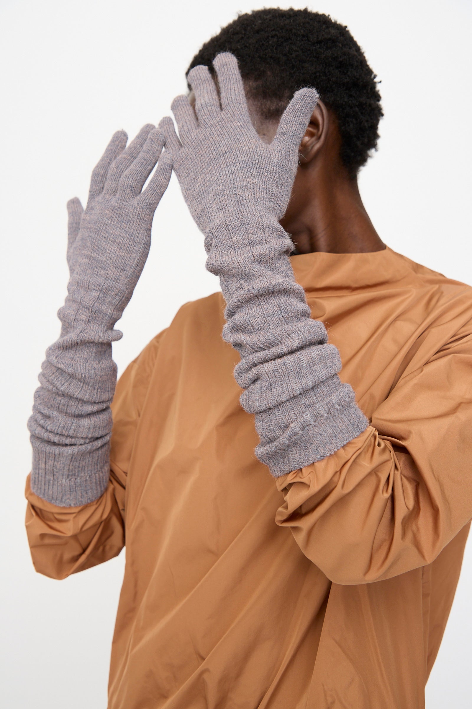 A person wearing a tan long-sleeve top and Rachel Comey Baalam Gloves in Dusty Mauve covers their face with gloved hands against a plain background.