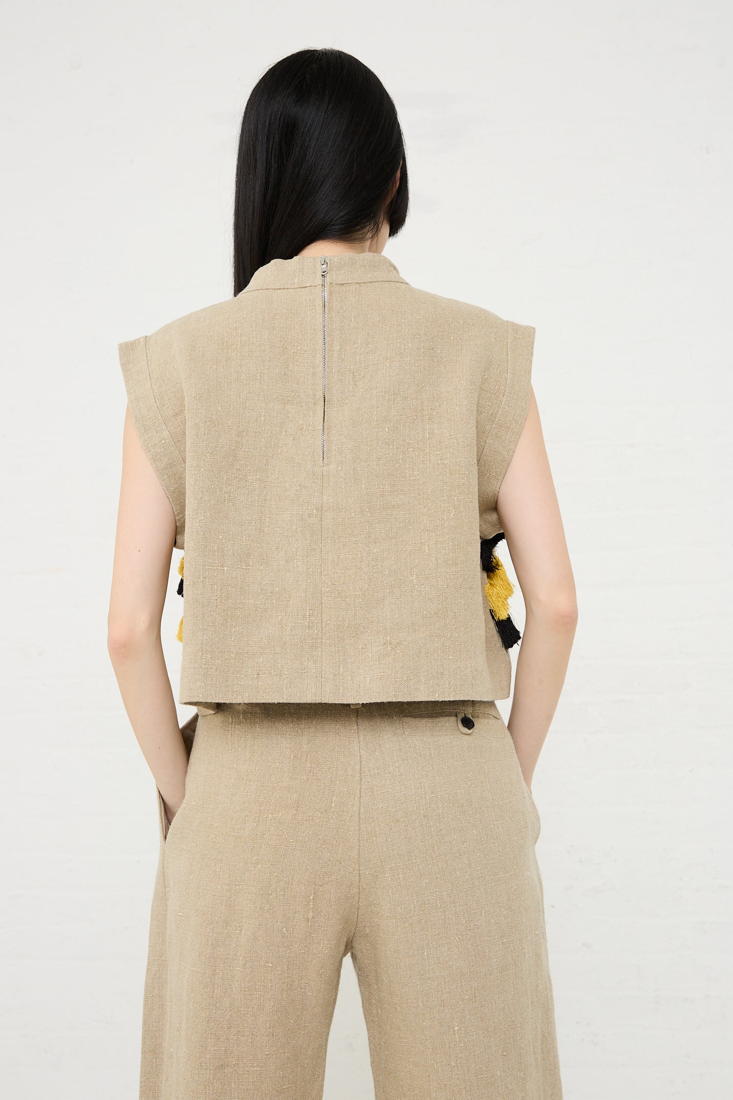 A woman seen from behind wearing a Rachel Comey Bacchus Top in Natural linen sleeveless crop top and matching trousers with abstract embroidery visible on the sides.