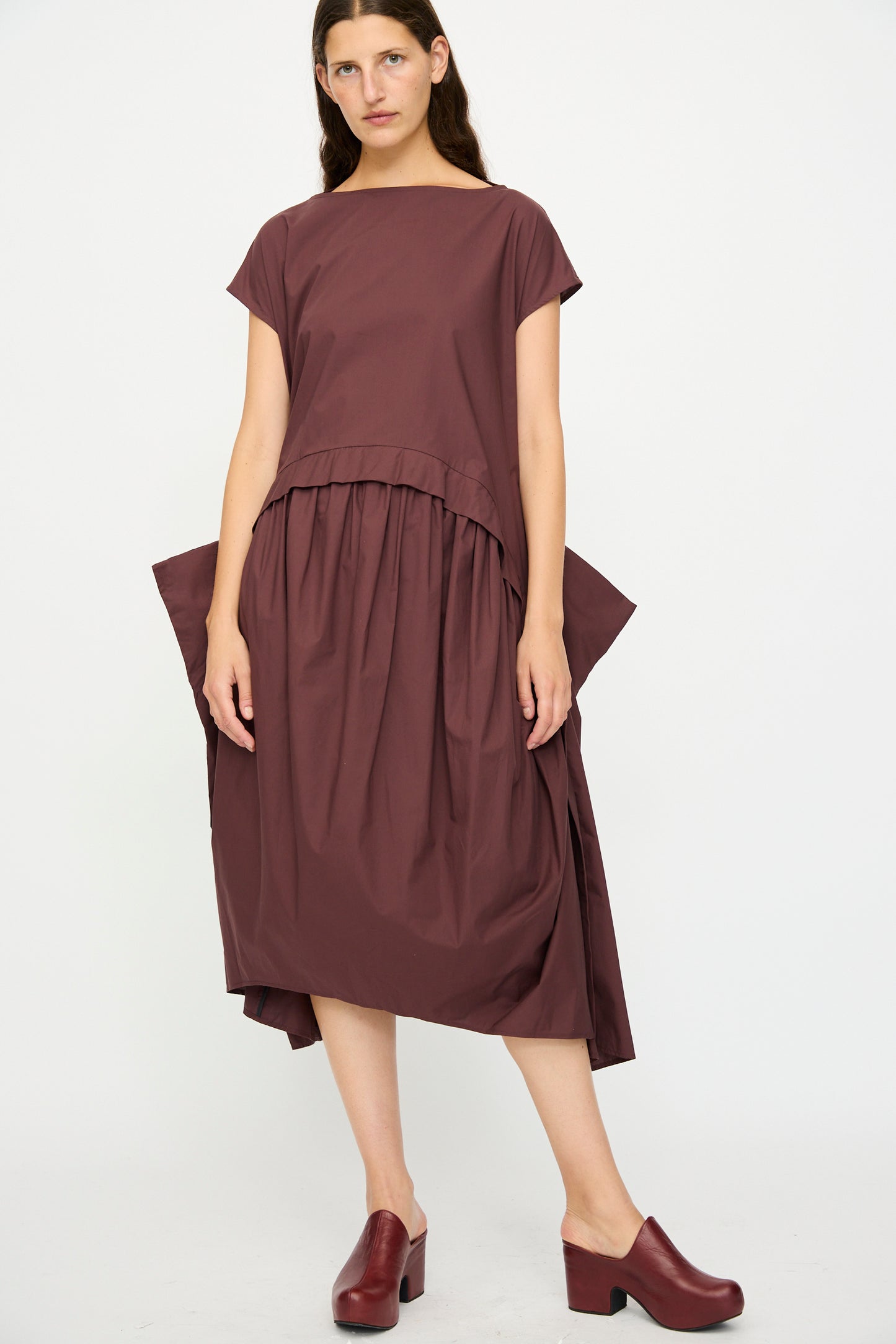 A woman stands against a plain background wearing the Rachel Comey Beau Dress in Merlot, an organic cotton midi with an asymmetrical hem, paired with matching burgundy heeled shoes.