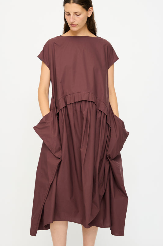 A woman wears the Rachel Comey Beau Dress in Merlot, a loose-fitting, short-sleeved tiered dress with large side pockets, standing against a plain white background.