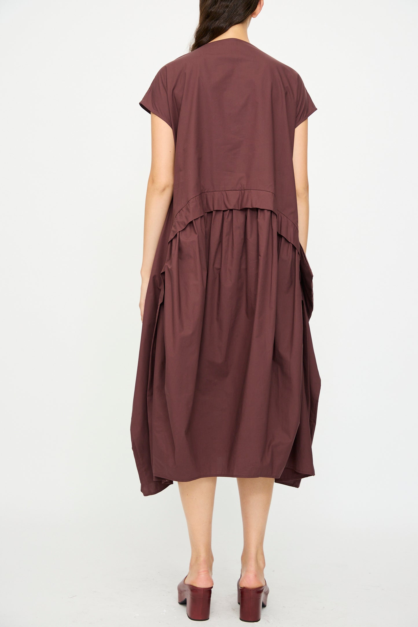 A person faces away, wearing the Rachel Comey Beau Dress in Merlot—an organic cotton, loose-fitting, short-sleeve midi dress with gathered details—paired with maroon platform sandals against a plain white background.