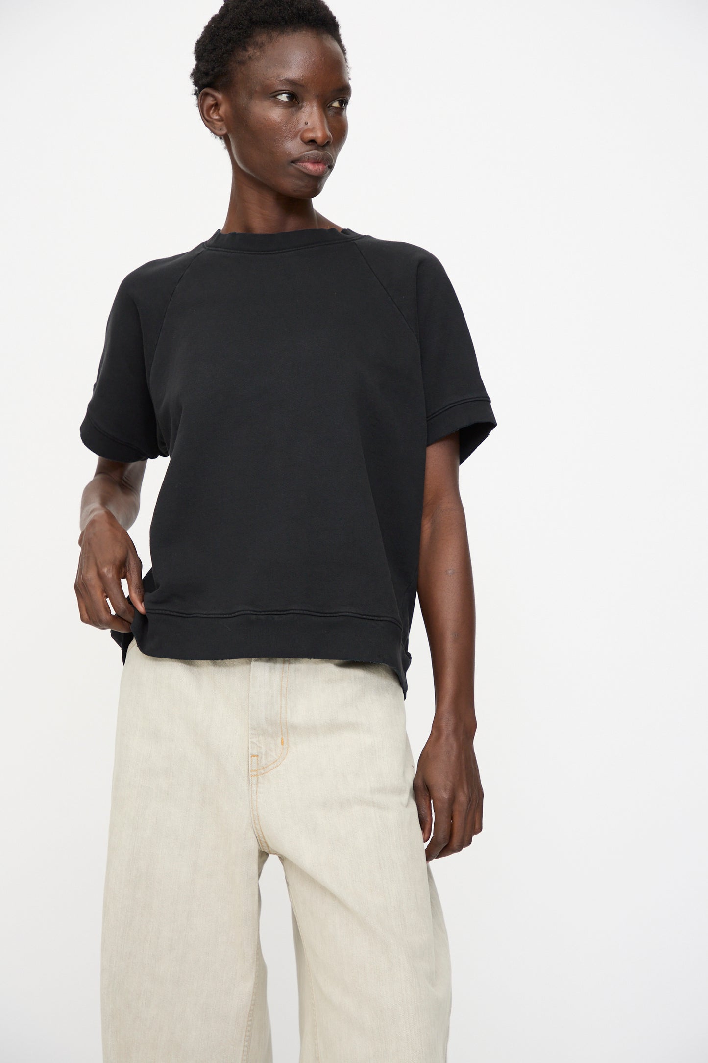 A person wears the Rachel Comey Billi Sweatshirt in Charcoal, a black 100% cotton crewneck short sleeve top, paired with light beige pants, standing against a plain light background and looking slightly to the side.
