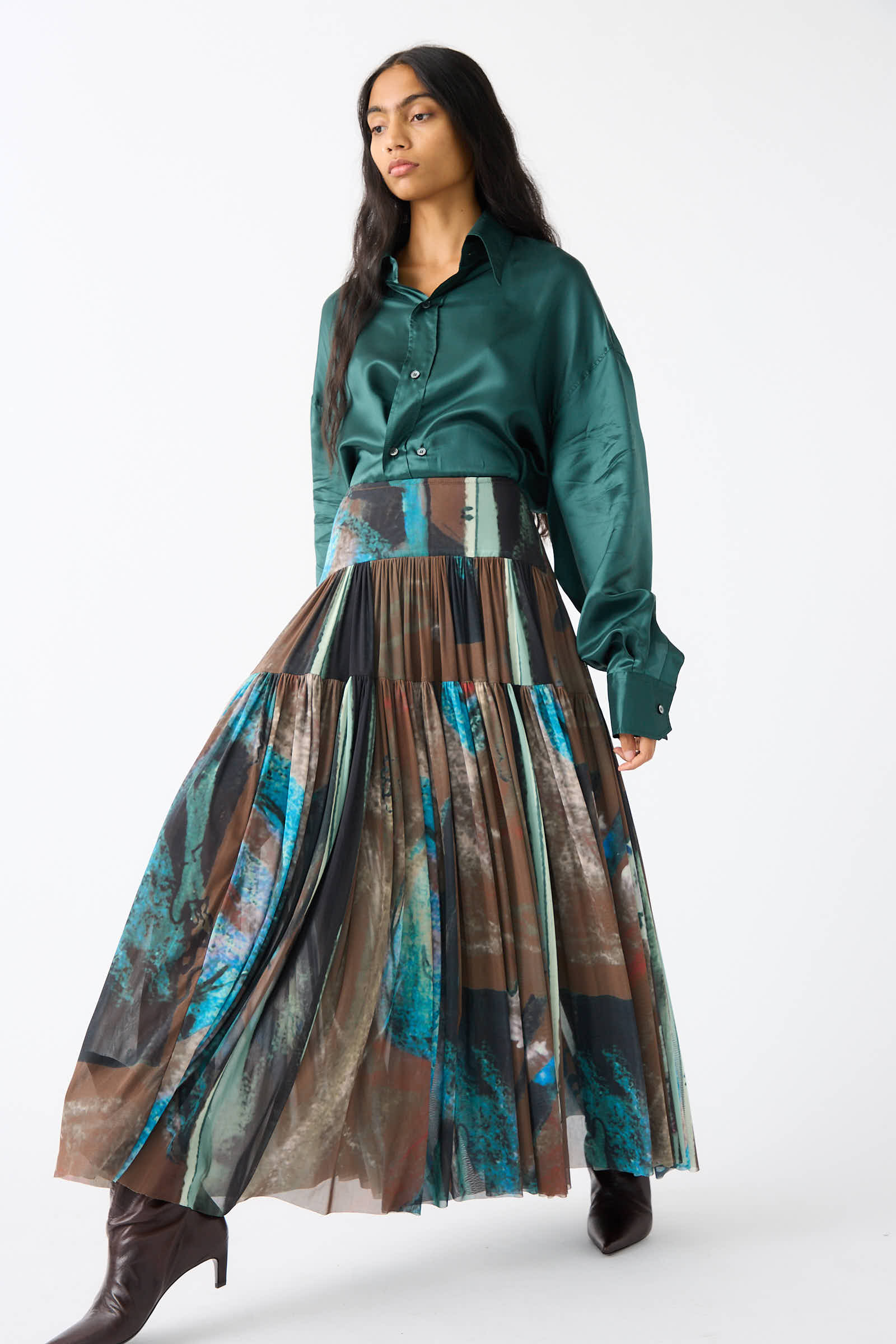 A person stands against a white background wearing a green satin long-sleeve shirt and Rachel Comey's Brush Print Mesh Flock Skirt in Brown, showcasing its colorful, abstract brush prints and pleated maxi design.