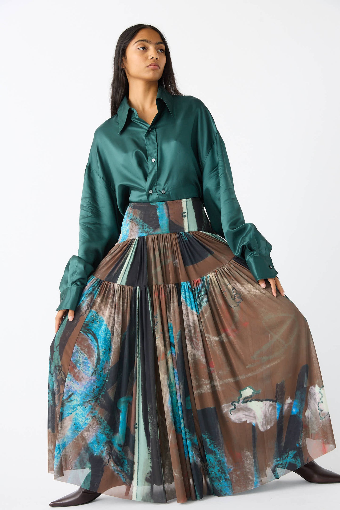 A person stands against a white background wearing a dark green long-sleeve shirt and the Brush Print Mesh Flock Skirt in Brown from Rachel Comey, which features a colorful, pleated maxi design with an abstract brush print.