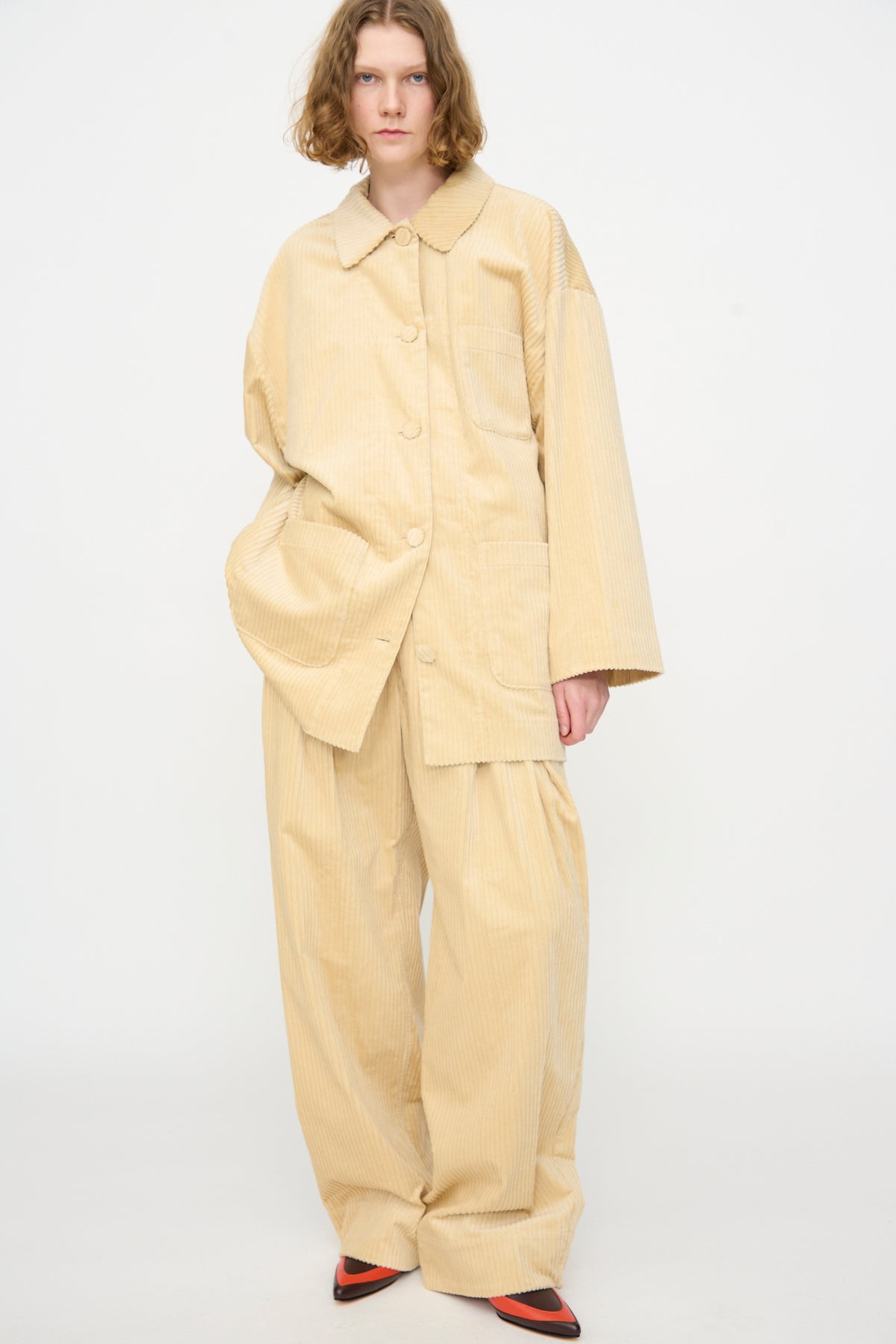 A person wearing the Rachel Comey Chore Jacket in Dune—an oversized beige cotton corduroy jacket—paired with matching wide-leg pants and black and red pointed shoes, stands against a plain white background.
