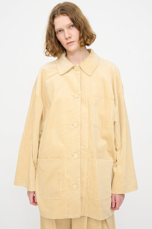 A person with shoulder-length light brown hair wears the Rachel Comey Chore Jacket in Dune—an oversized beige cotton corduroy style featuring large buttons and three front pockets—while standing against a plain white background.