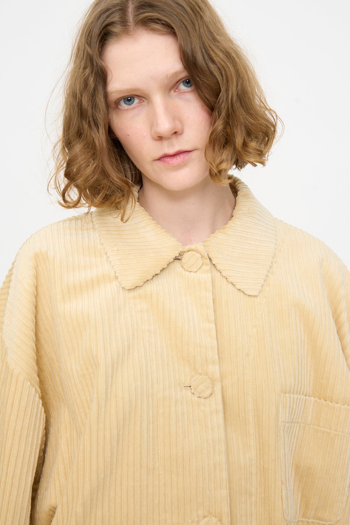 A person with wavy, shoulder-length light brown hair wears the Rachel Comey Chore Jacket in Dune—an oversized, pale beige cotton corduroy piece featuring a wide collar and front pocket—standing against a plain light background.