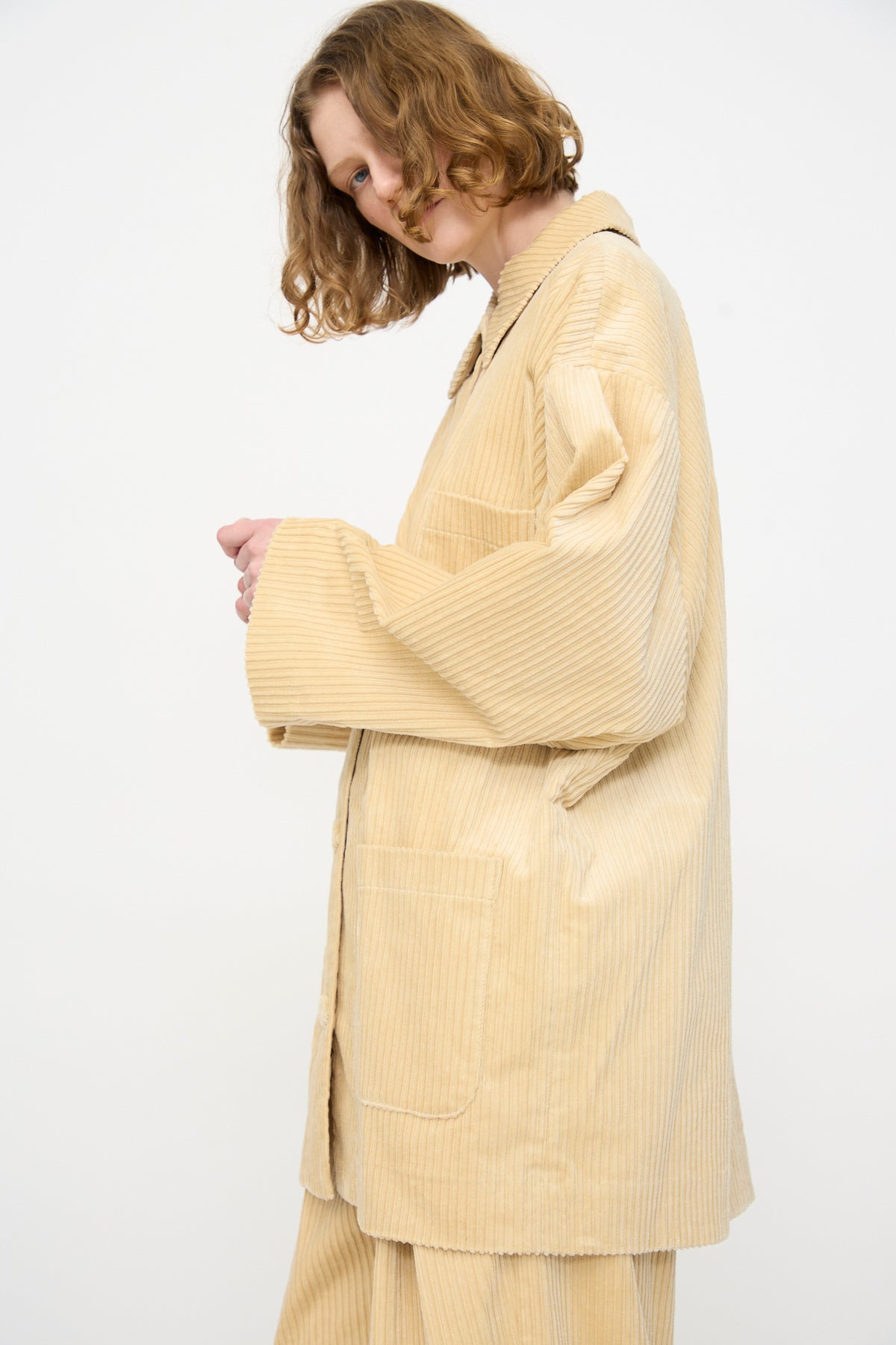 Person with wavy, shoulder-length hair wearing the Rachel Comey Chore Jacket in Dune with matching pants, standing against a plain white background.